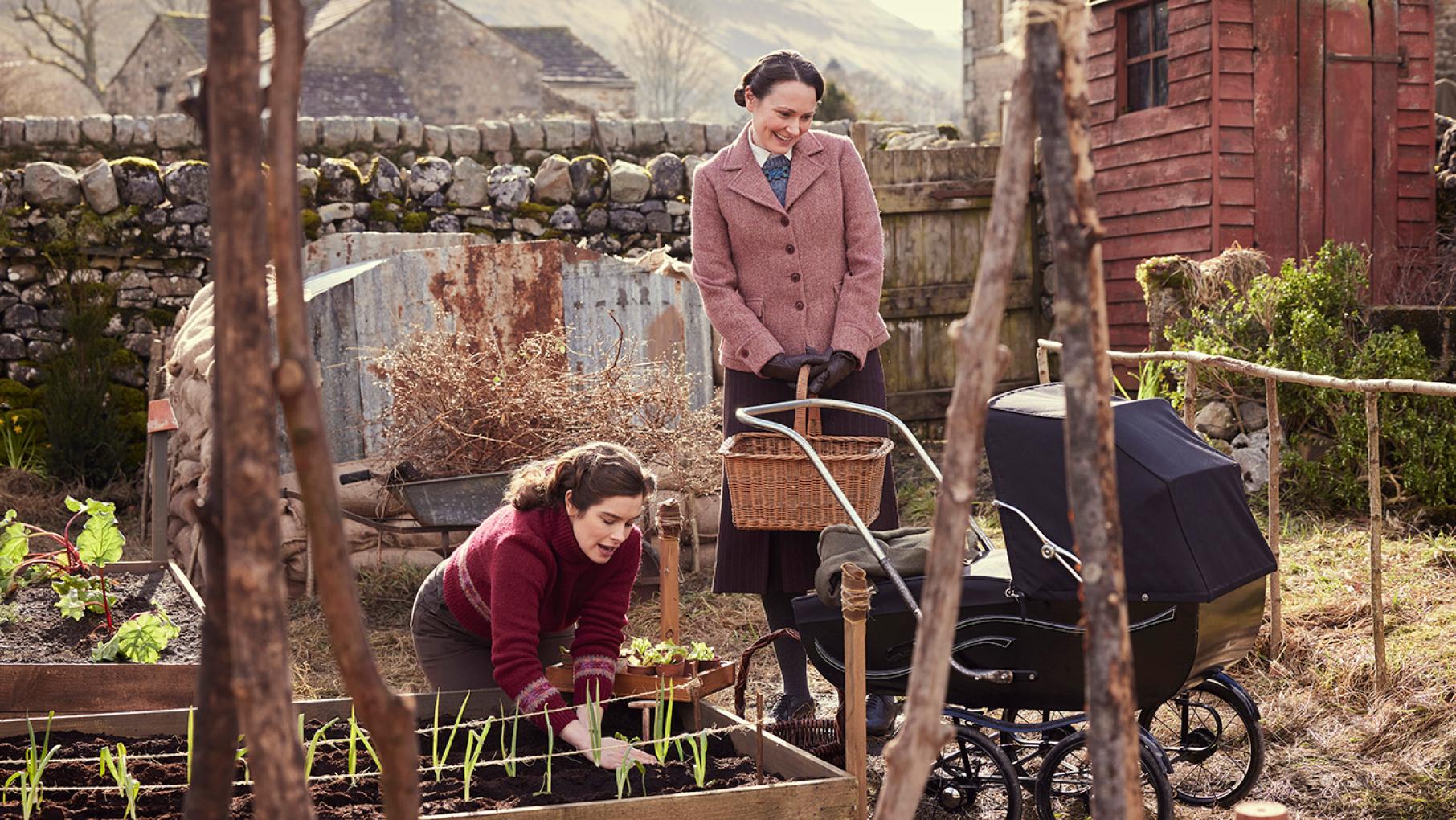 Darrowby is contributing to the war effort with victory gardens, knitting drives, and its men in the military. Credit: Helen Williams for Playground Entertainment and Masterpiece Helen works in a raised garden bed while Mrs. Hall stands by and smiles