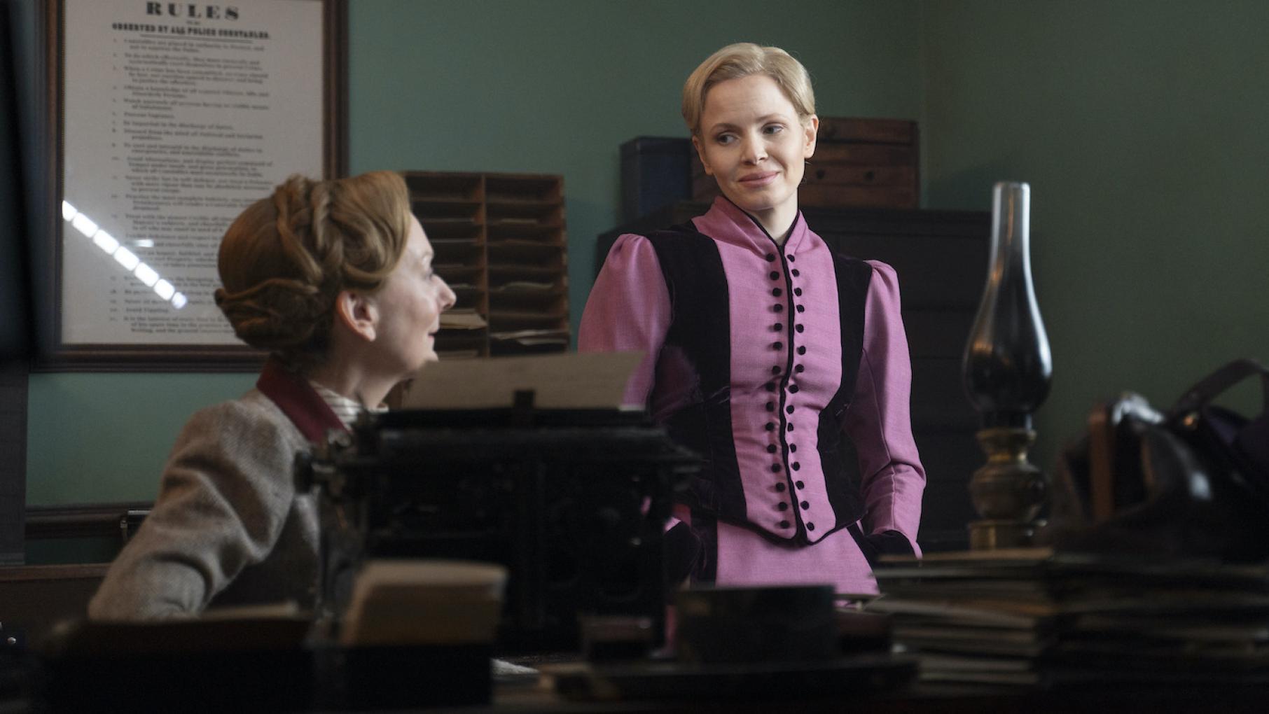 Eliza stands and looks at a seated Ivy