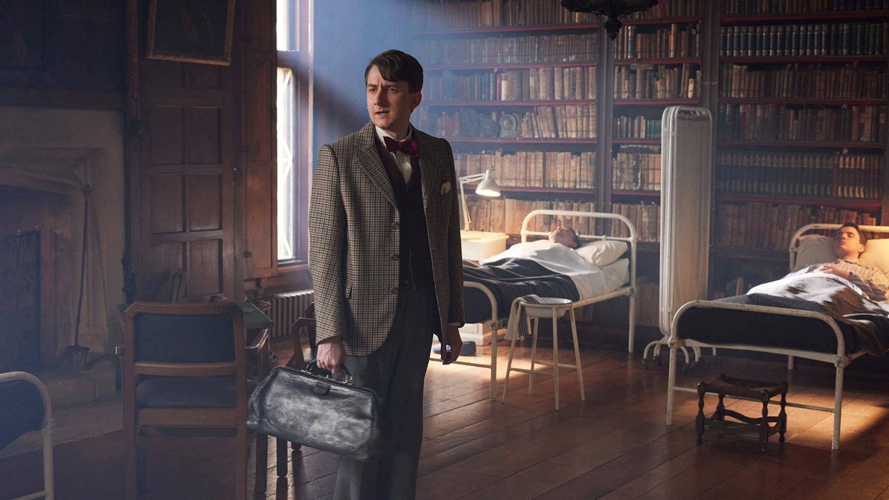 Richard stands holding a bag in a library with two men lying in cots behind him