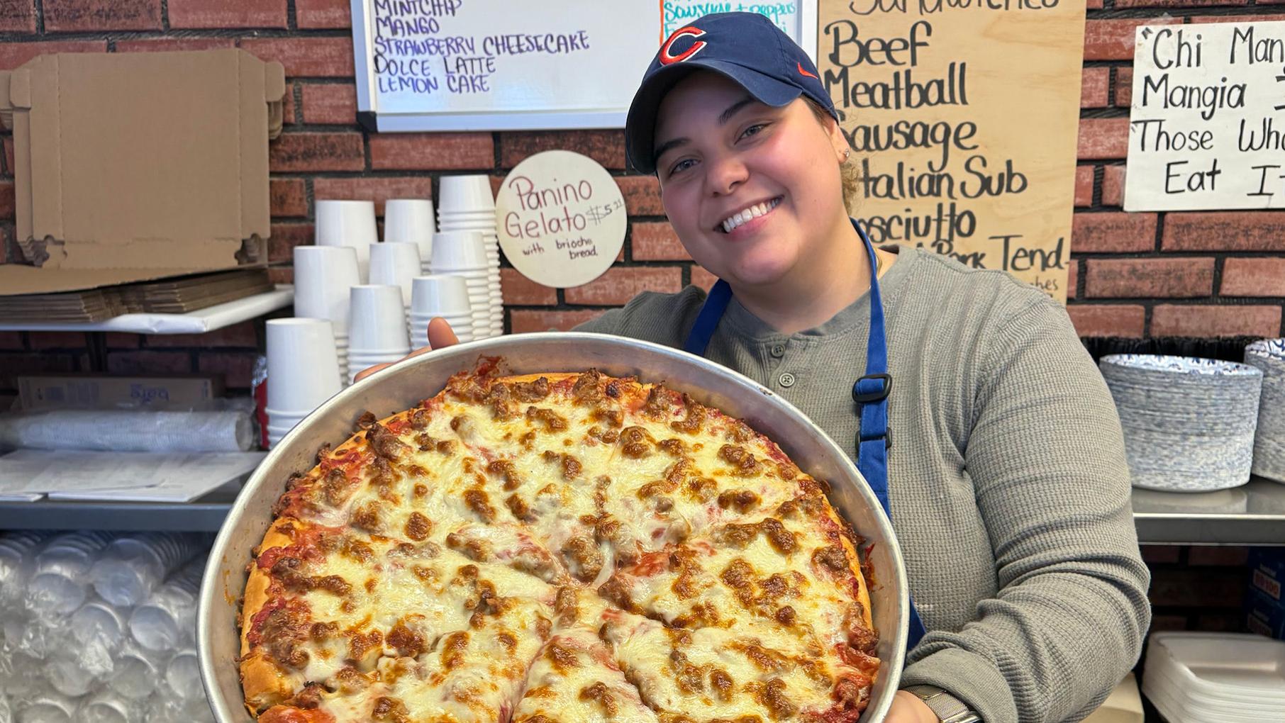 Freddy's has been forging bonds with customers in Cicero thanks to their pizza, Italian dishes, and hospitality for decades. Credit: David Hammond for WTTW A woman in a Cubs hat smiles and holds up a large circular pan of sausage pizza