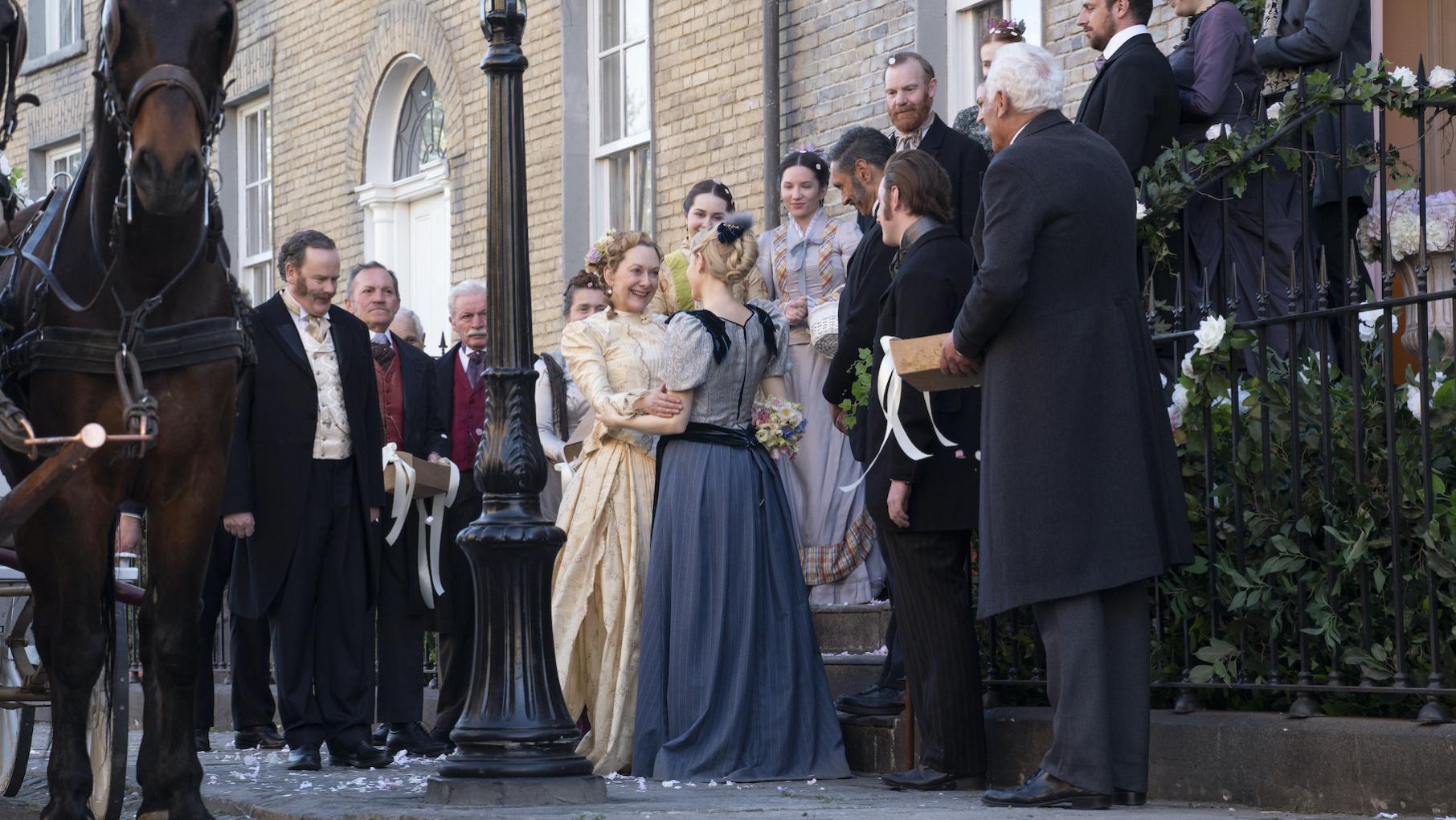 Ivy and Eliza look at each other outside their house as wedding guests look on and horses wait