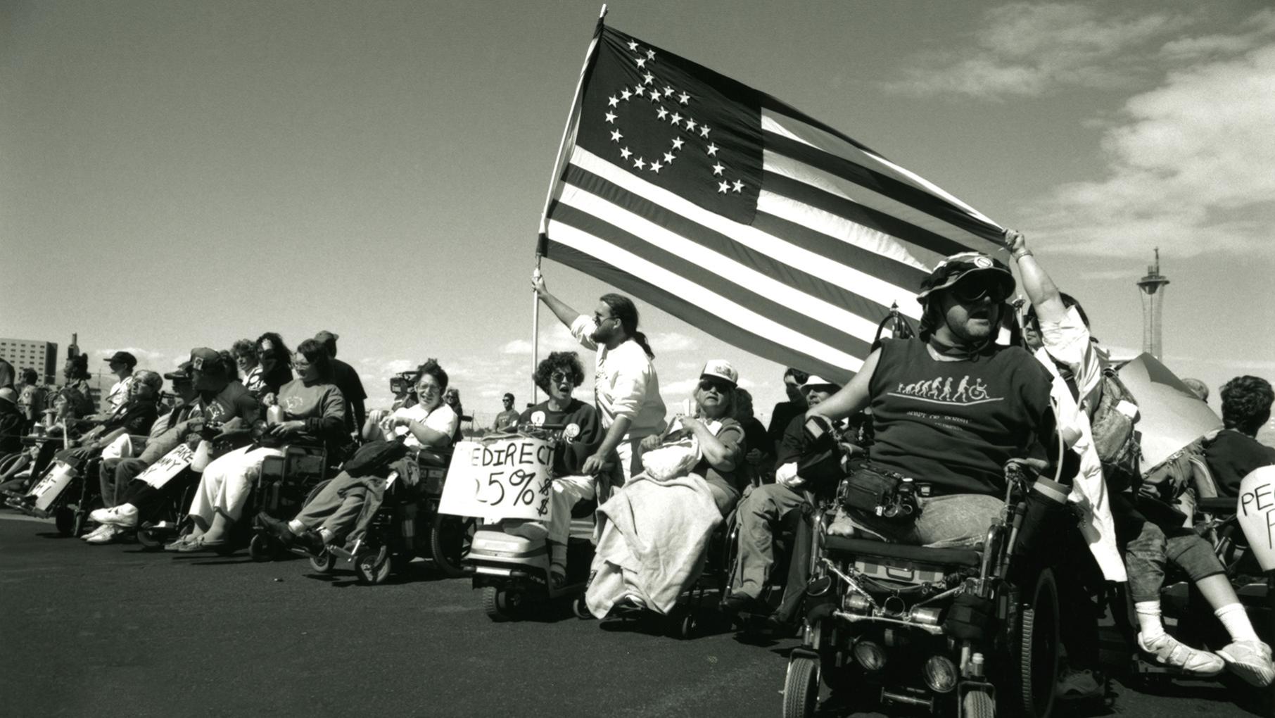ADAPT (American Disabled for Accessible Public Transit) activists protest in Las Vegas. Credit: Tom Olin A line of protesting people in wheelchairs under an American flag with a wheelchair icon