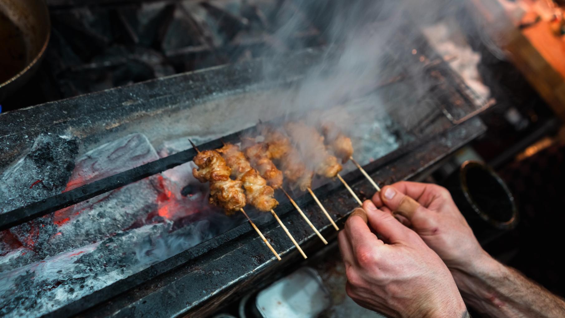 Hands turn small wooden skewers of chicken over charcoal in a Japanese yakitori set-up
