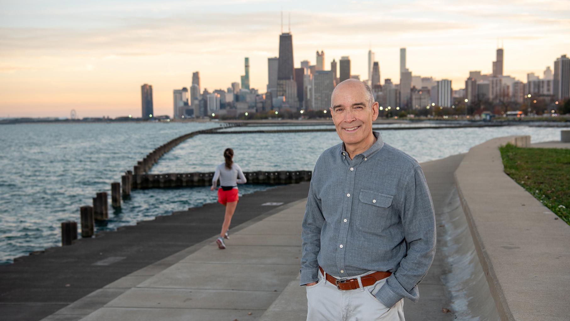 Geoffrey Baer smiles in front of Chicago's skyline and a woman running on the lakeshore