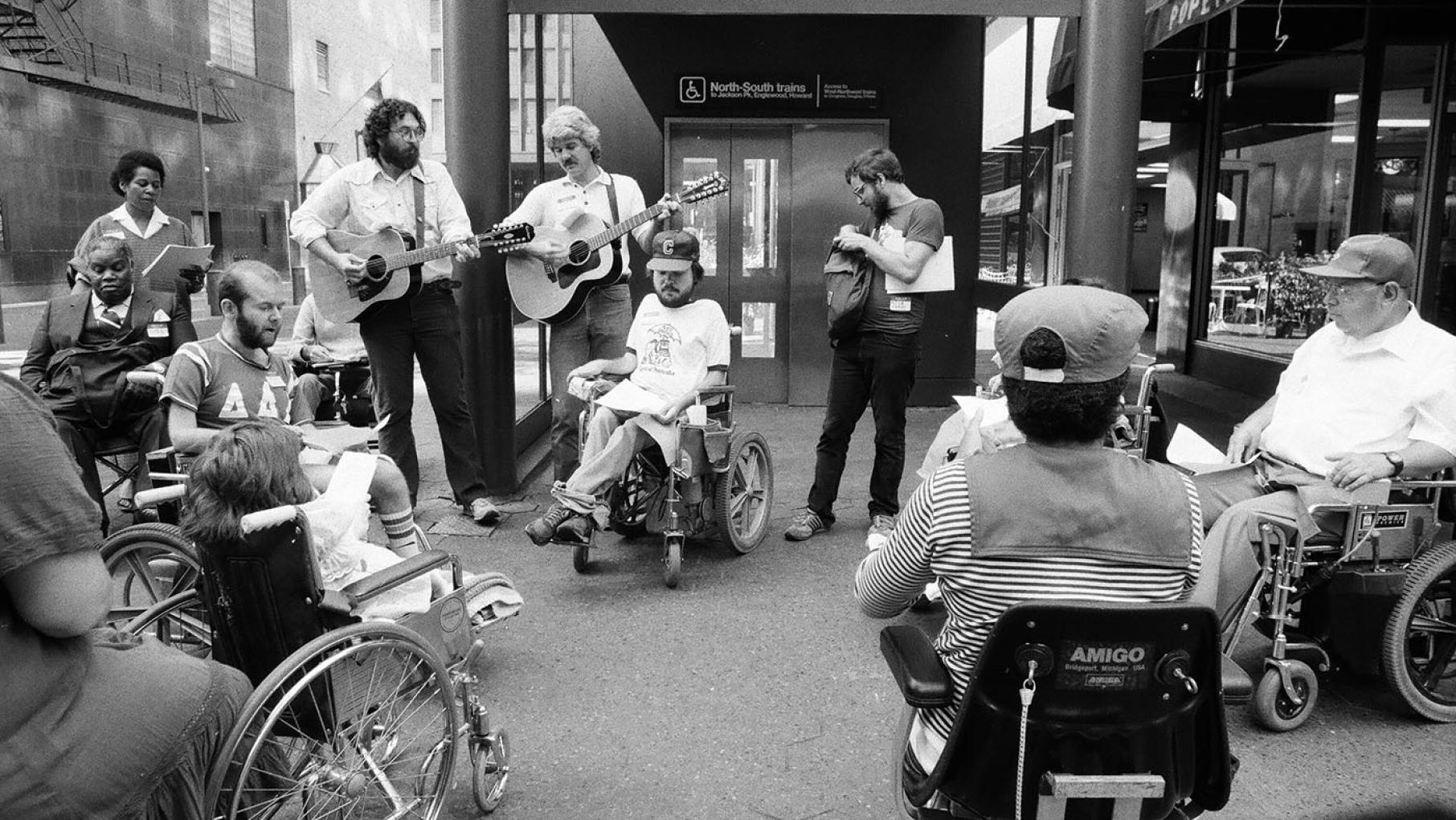 A black and white image depicts people in wheelchairs outside an entrance to a CTA station in downtown Chicago.