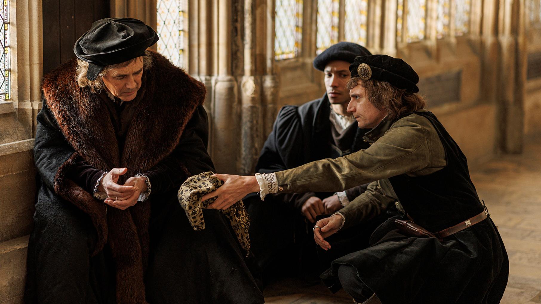 Cromwell sits against a wall as Richard Riche reaches out a hand and Christophe squats behind