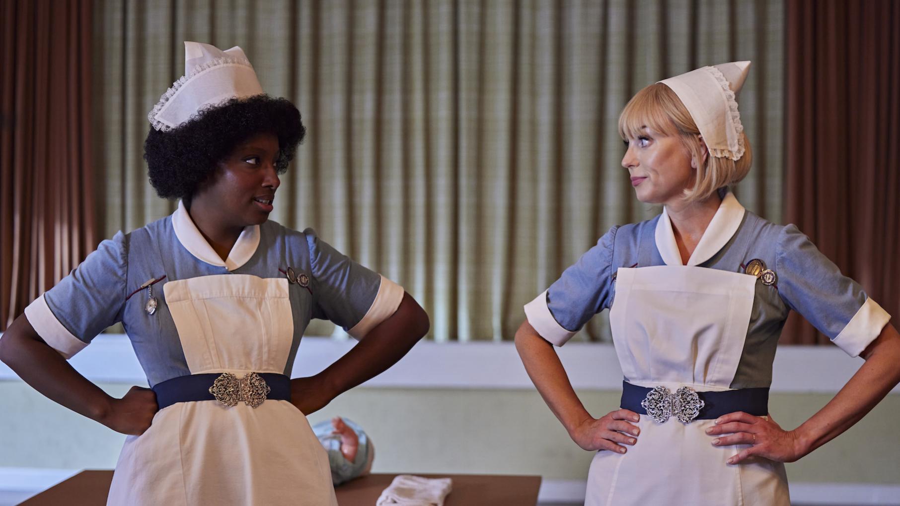 Joyce and Trixie stand with their hands on their hips in front of a stage in their nurses' uniforms
