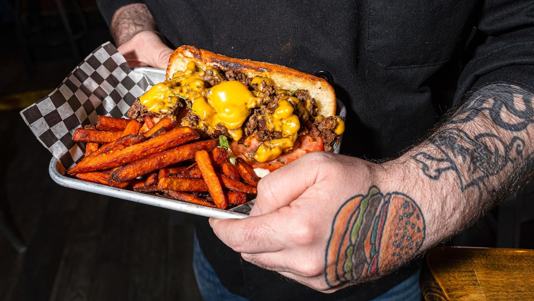 A sandwich with a cheese sauce and sweet potato fries arranged on a try carried by a person with a hamburger tattoo on the back of his hand.