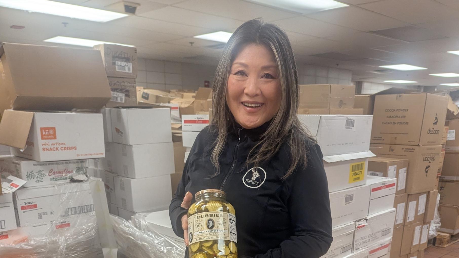 Jiwon McCartney helped found Fight2Feed to distribute food that would otherwise go to waste to those in need. Credit: Lisa Futterman for WTTW A woman holds a jar of pickles and smiles in a room full of boxes