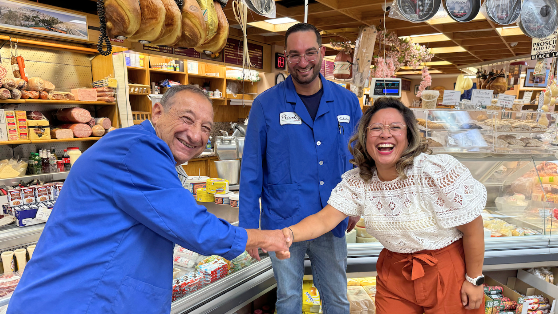 A man in a blue coat smiles while he shakes the hand of a woman who is also smiling, in front of another smiling man, in a grocery store
