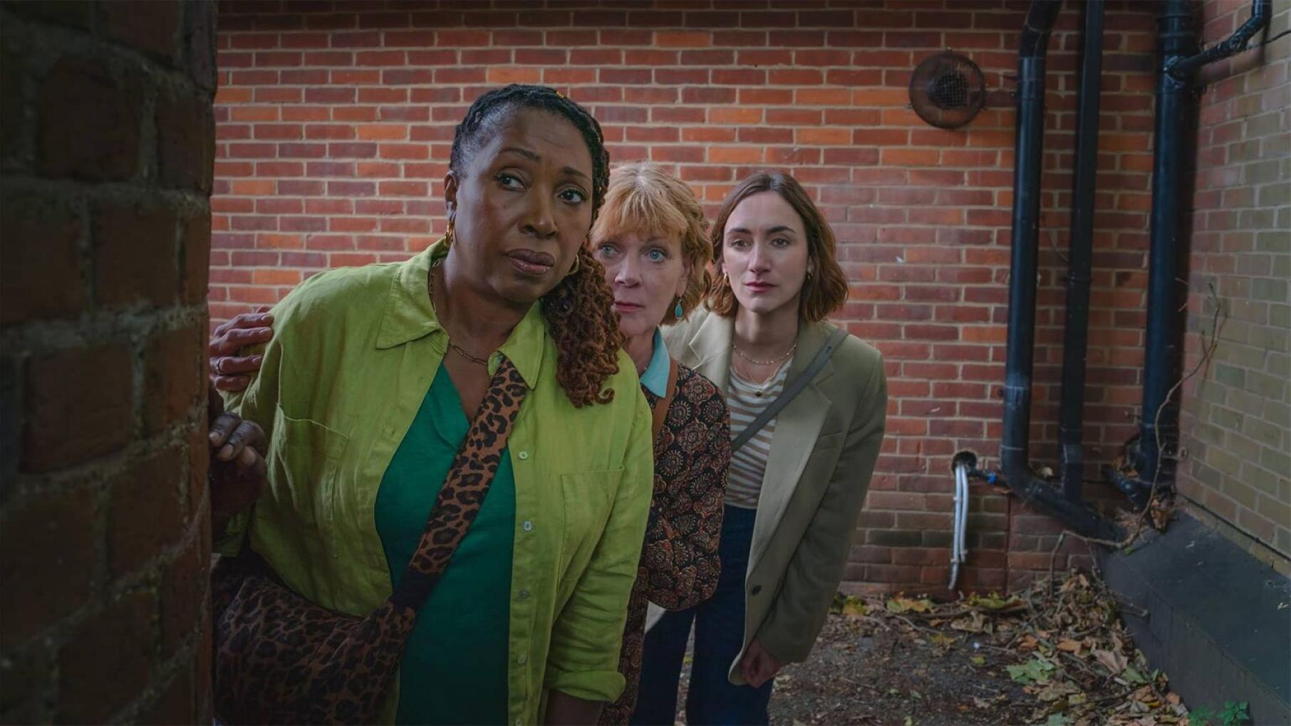 Suzie, Judith, and Becks are back to solve more crimes in 'The Marlow Murder Club.' Credit: Masterpiece Three women peer around a corner