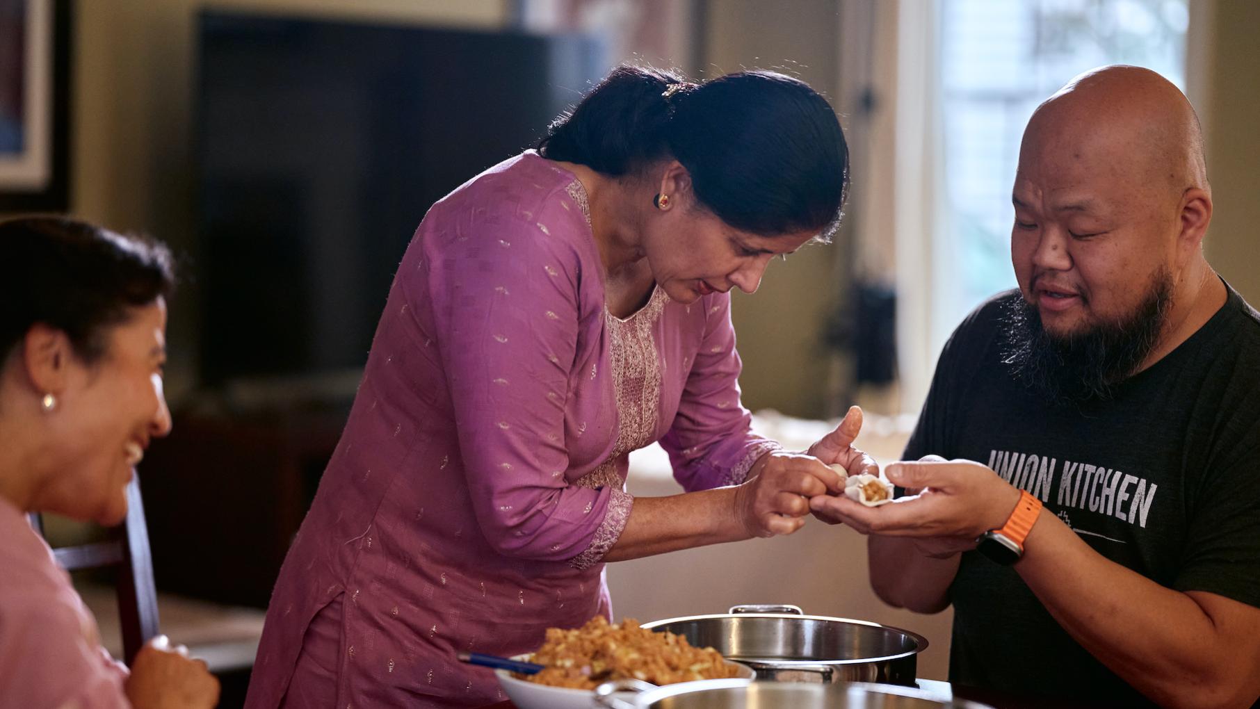 In the food series 'Relish,' chef Yia Vang learns to make dishes like Nepali momos while hearing the personal stories and cultural traditions behind them. Credit: Uche Iroegbu for Twin Cities PBS A woman helps fold a dumpling in a man's hand at a table while another woman watches
