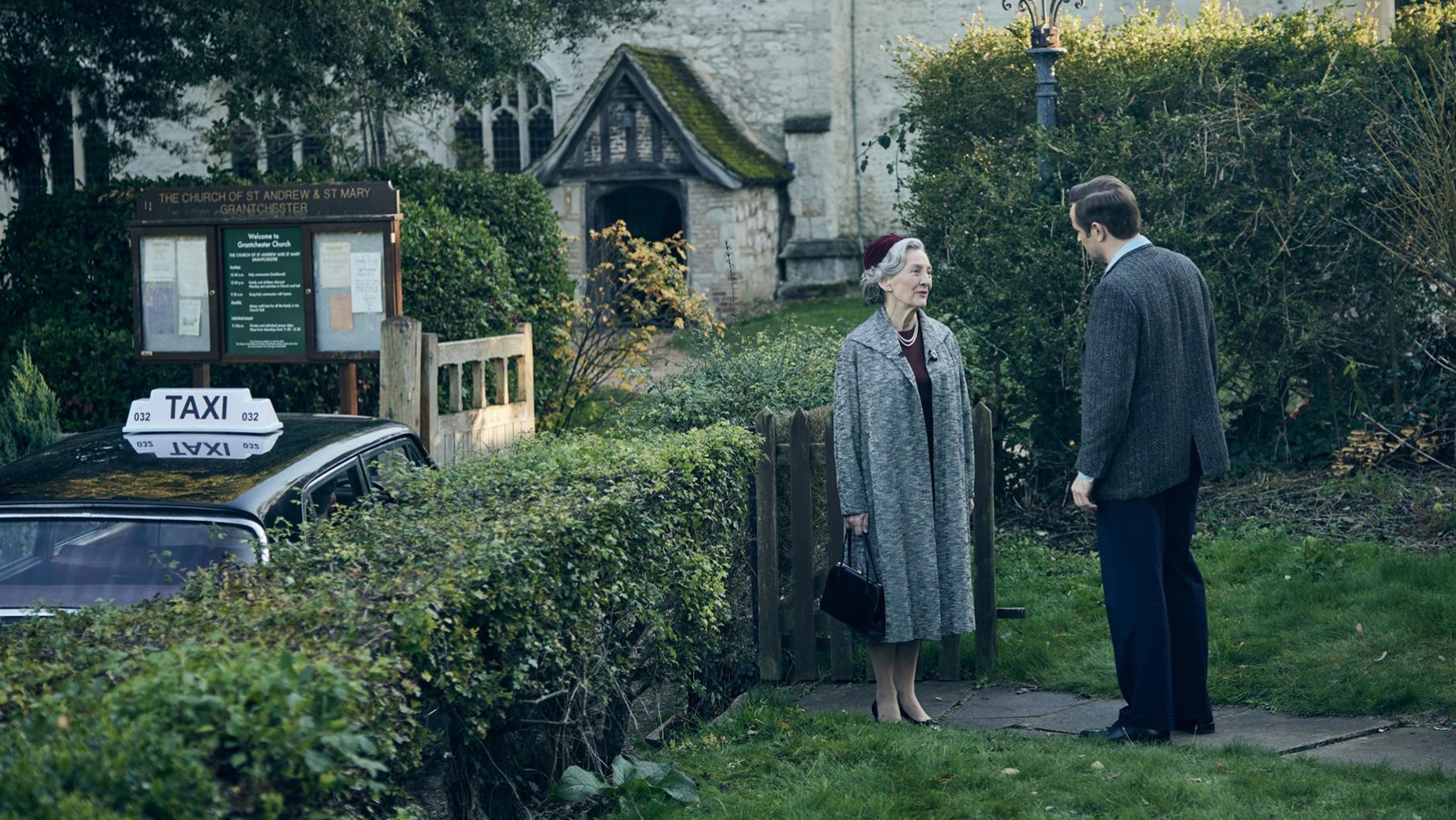 Daniel stands talking to his mother next to a hedge in front of the church