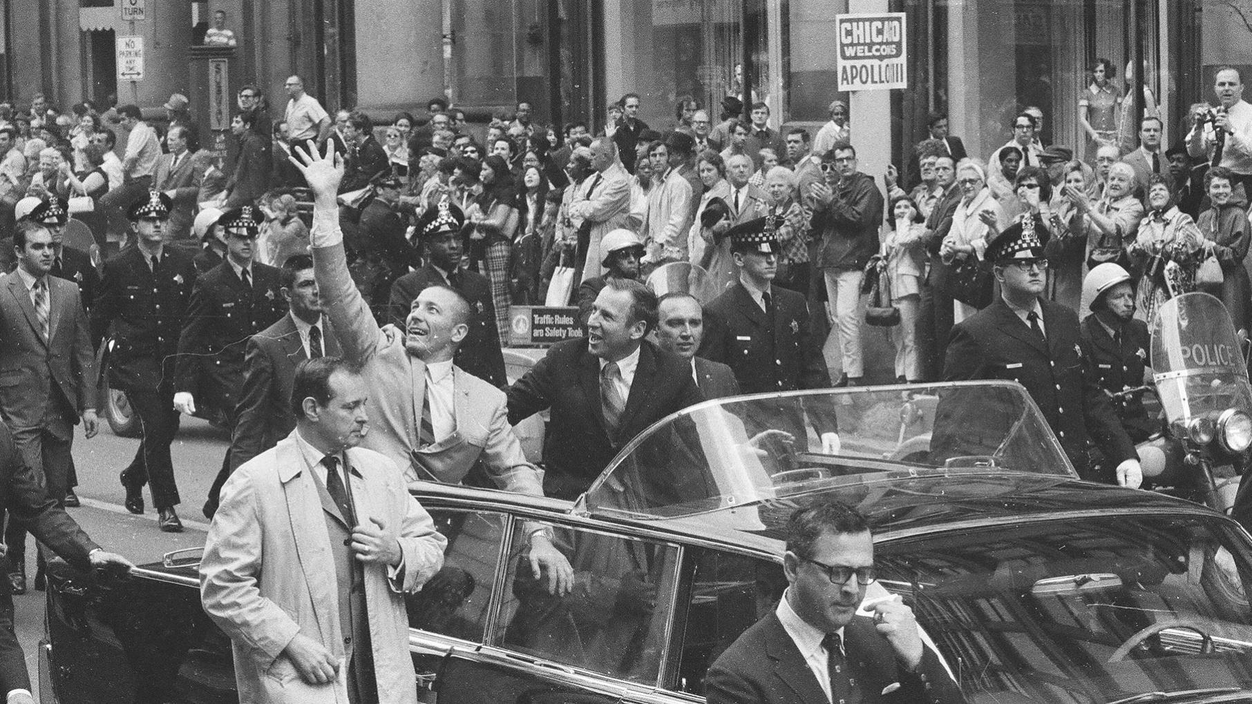 Apollo 13 astronauts John Swigert, left, and James Lovell sit in an open-top car at a parade in Chicago’s Loop in a black and white image.