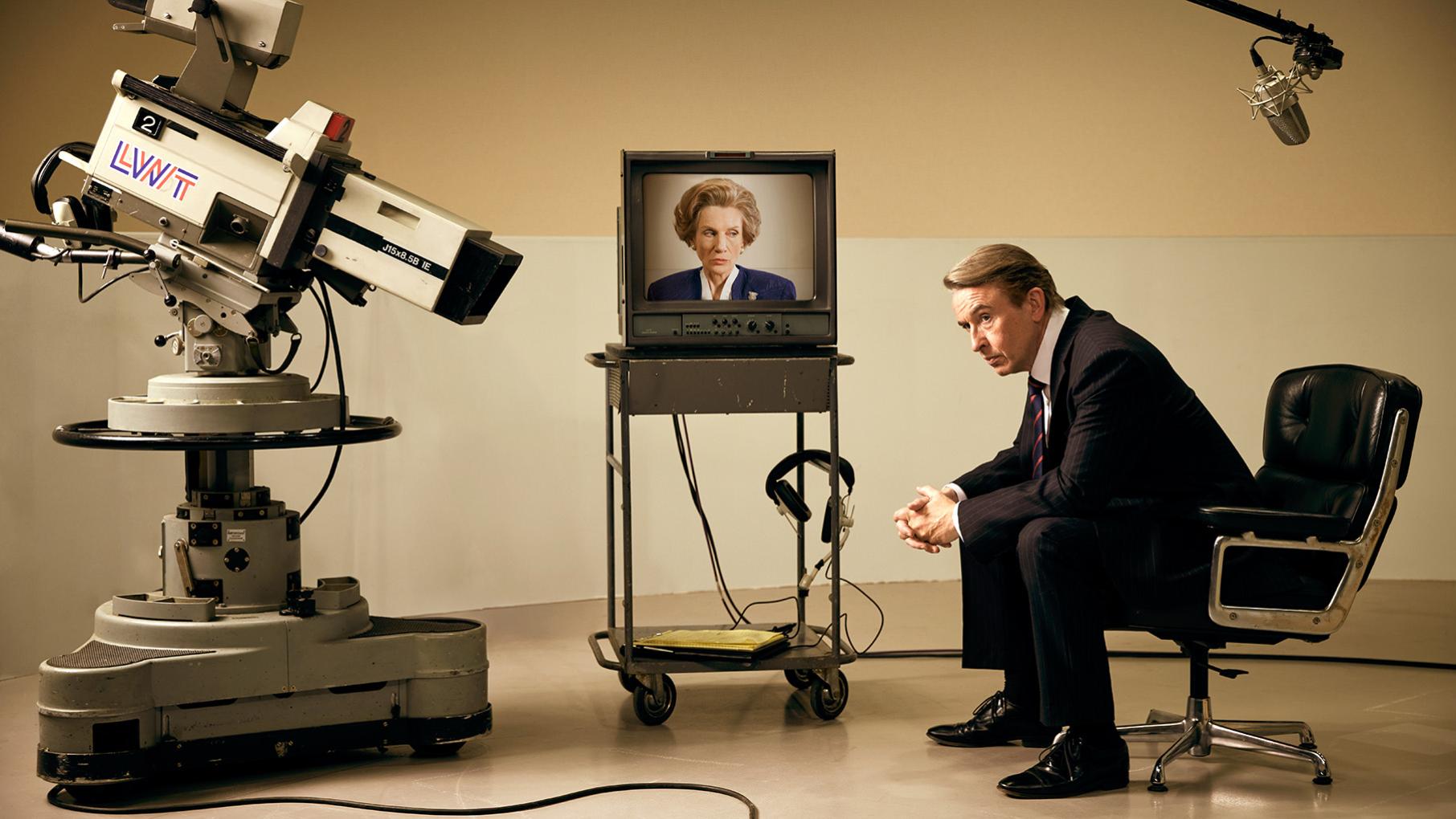 Brian Walden sits hunched in a chair on a TV set in front of a TV showing Margaret Thatcher, with a camera pointing at him