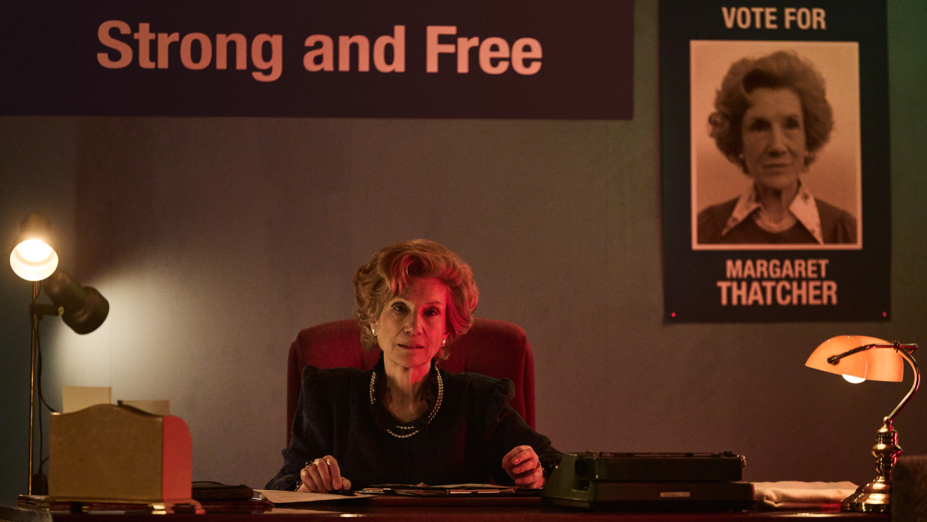 Margaret Thatcher sits at a desk in a dark campaign office in front of a poster of herself