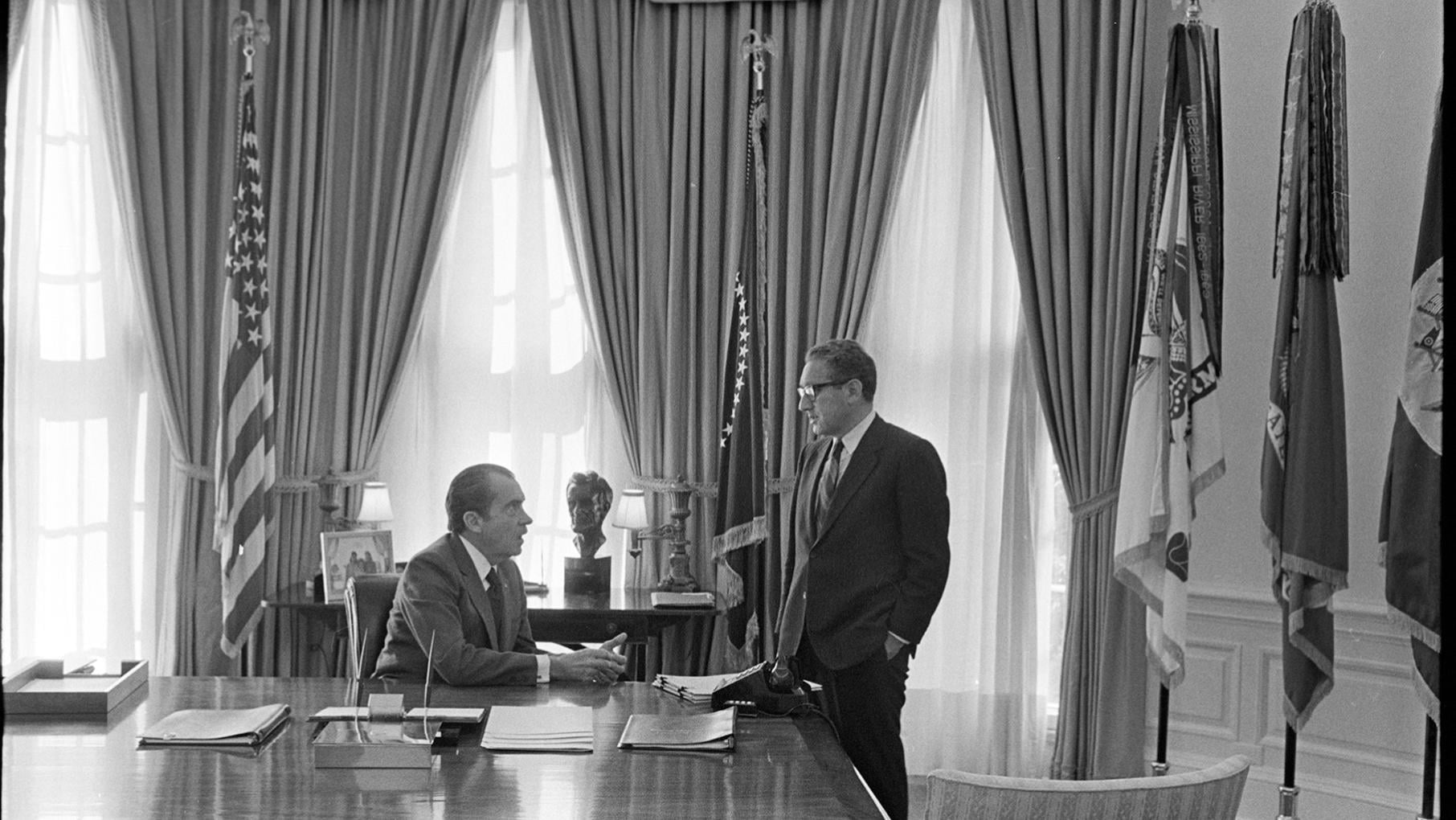 President Richard Nixon sits behind a desk while Henry Kissinger stands next to him in the Oval Office in a black and white photo