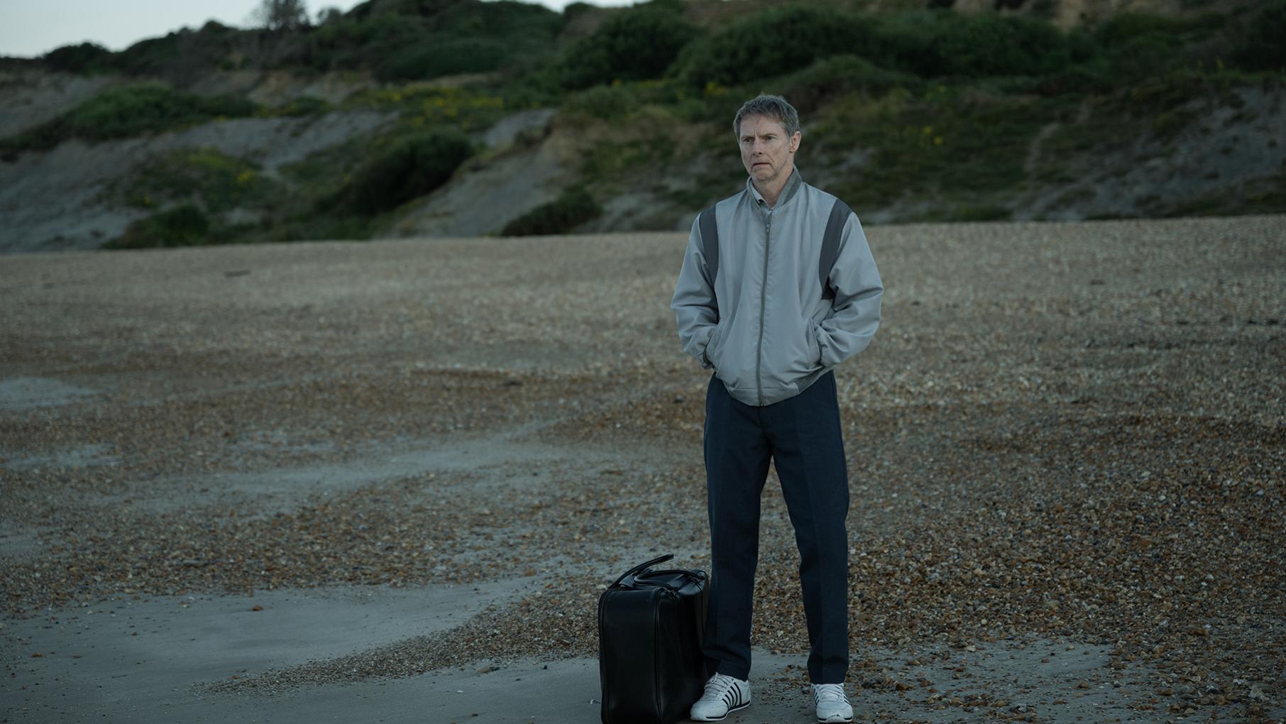 Gordon stands with his hands in his pocket on a beach looking out to sea with a suitcase next to him on the ground