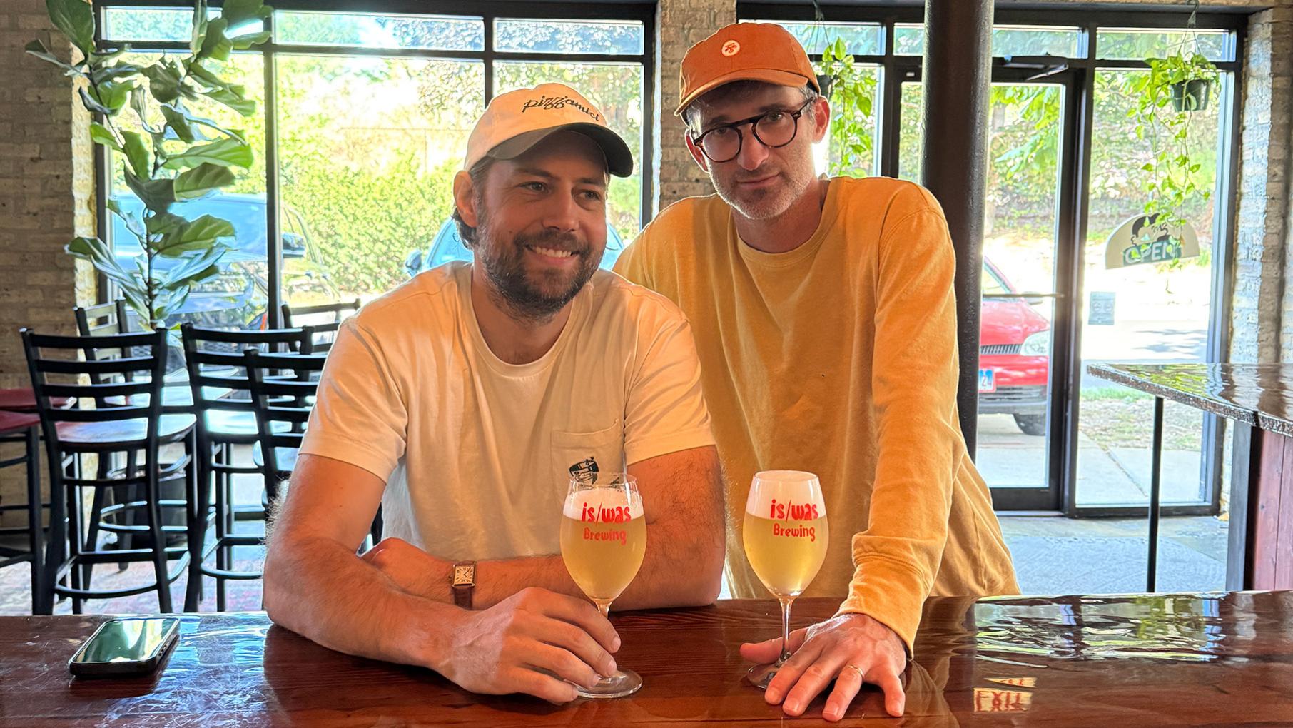 The two hosts of Joiners at a bar with beers pose for a photo