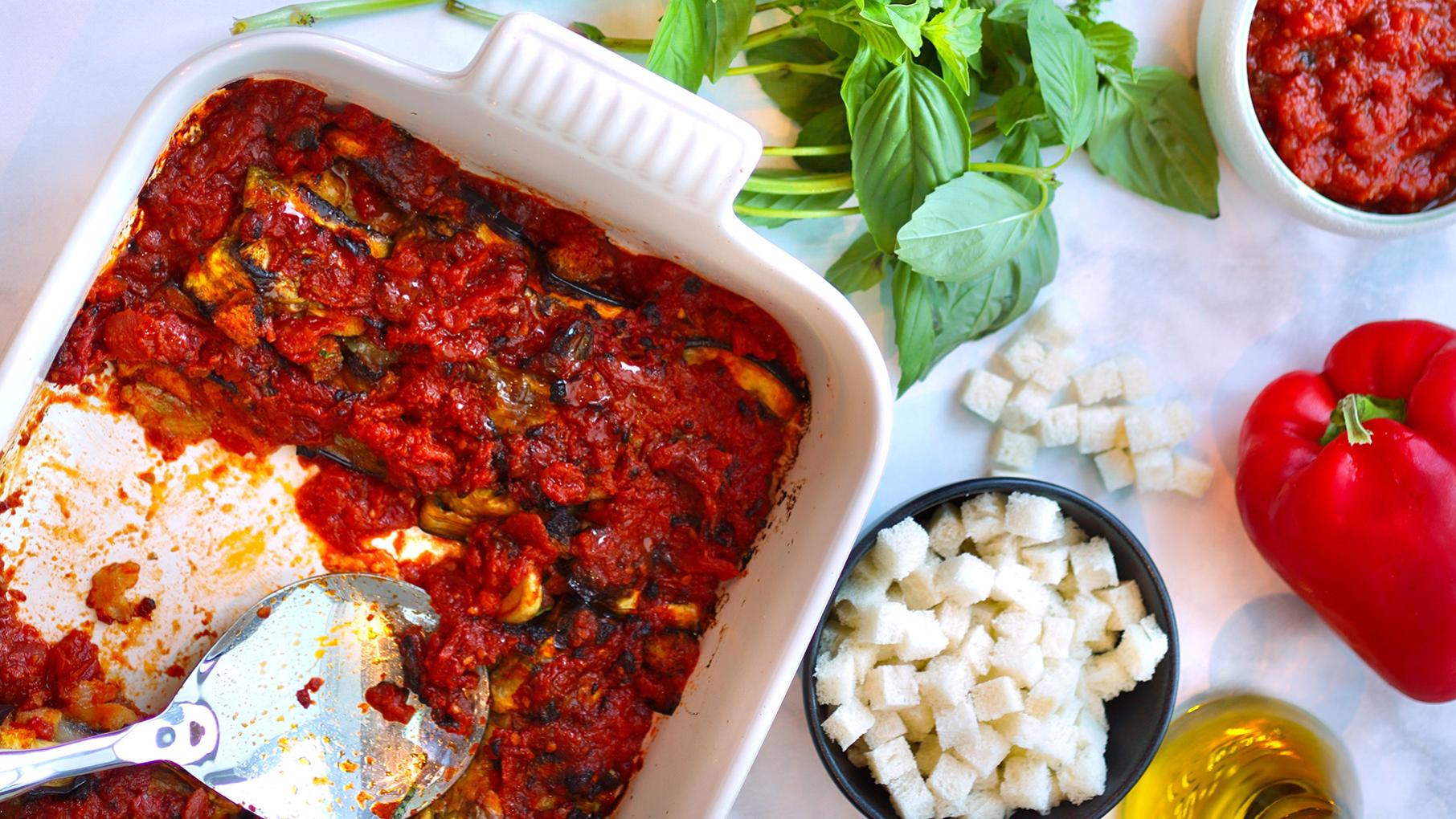 A baking dish of eggplant parmesan next to fresh basil, bread cubes, tomato sauce, and a red pepper