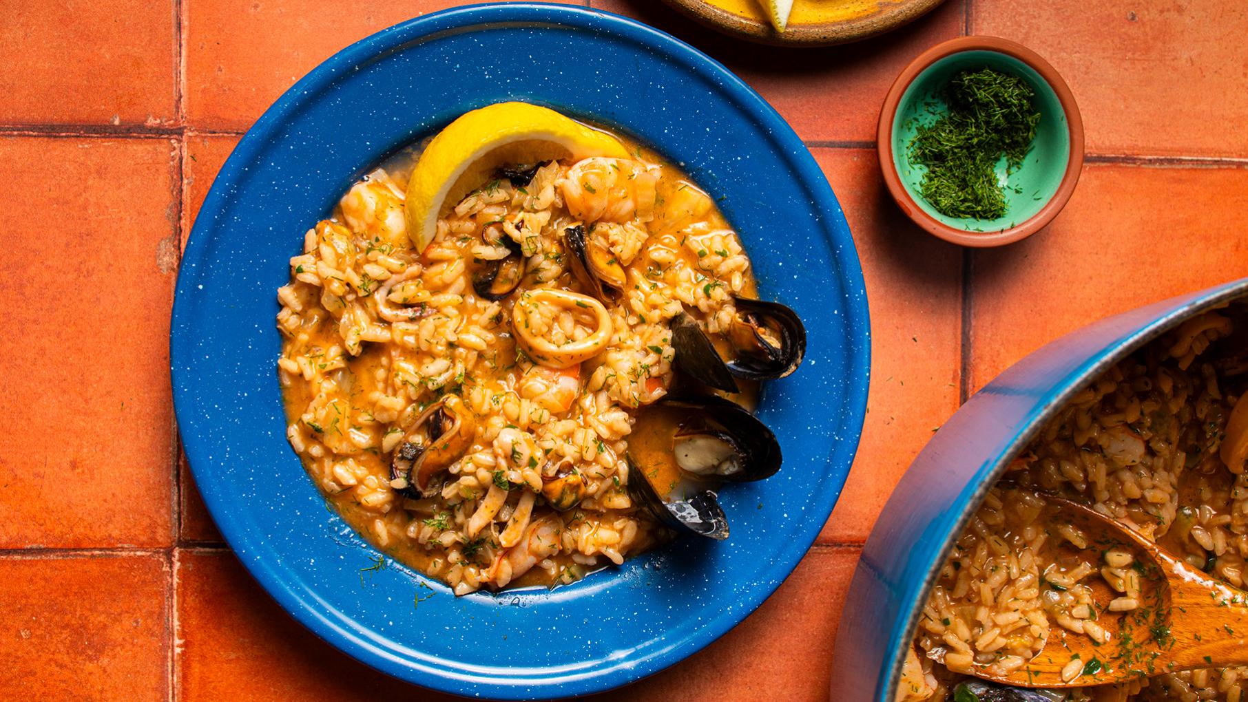 A blue bowl filled with seafood risotto next to a small bowl of herbs and a pot of more risotto