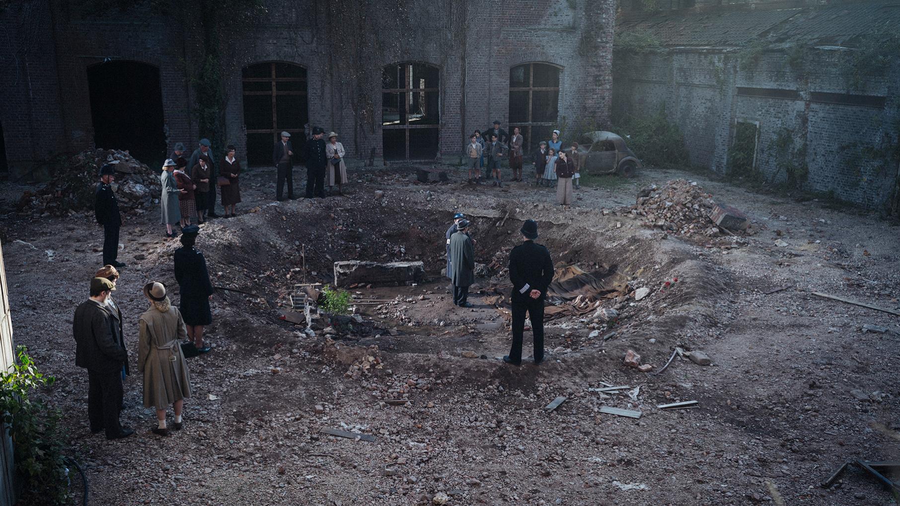 A crowd of people stand in and around a bomb crater in front of an abandoned building