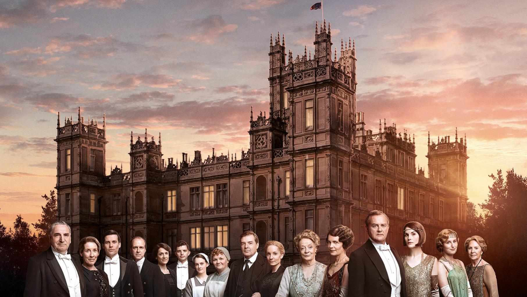 The caste of Downton Abbey in front of the abbey itself at sunset