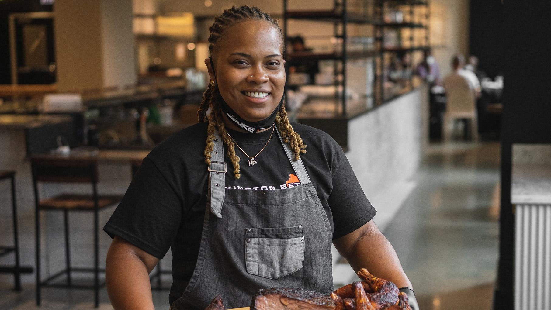 Dominque Leach smiles as she holds a platter of barbecue in an apron