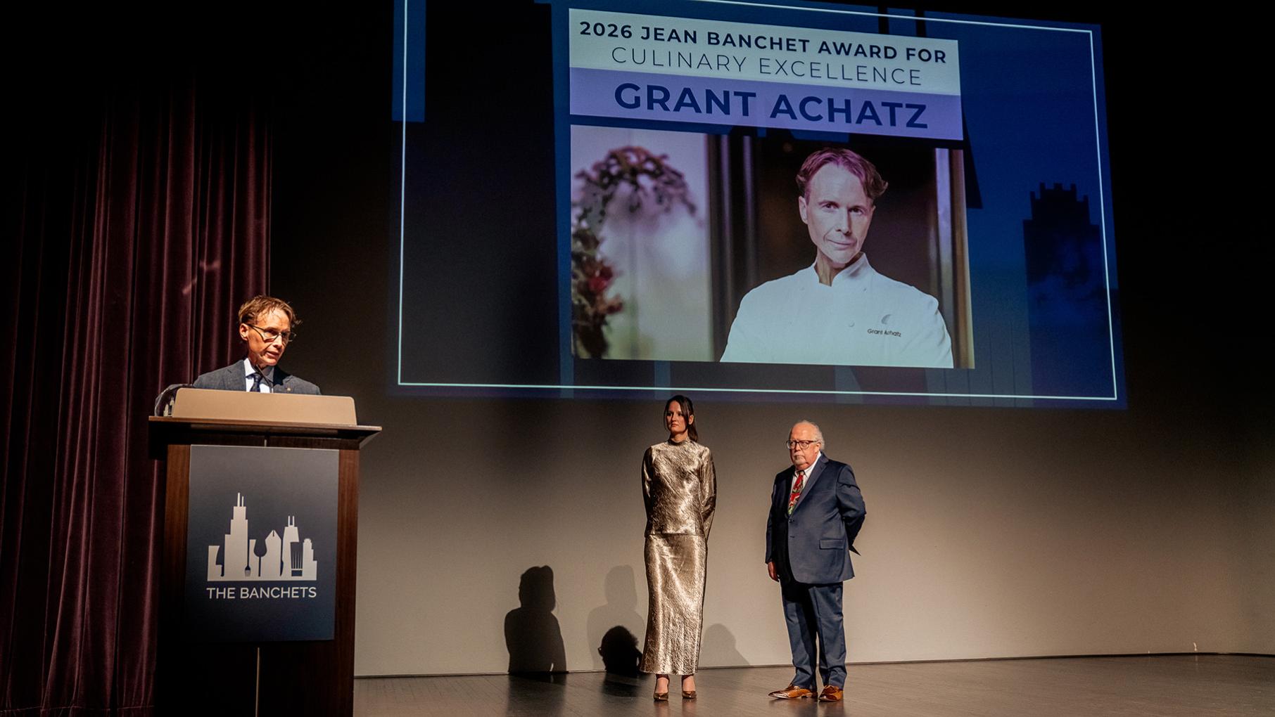 Grant Achatz stands at a podium on a stage near a screen with his photo and name and two people standing underneath it