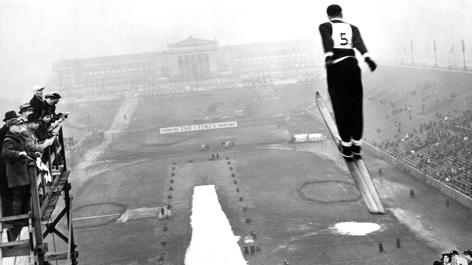 A black and white image of a ski jumper mid-air with people standing on a platform to the left.