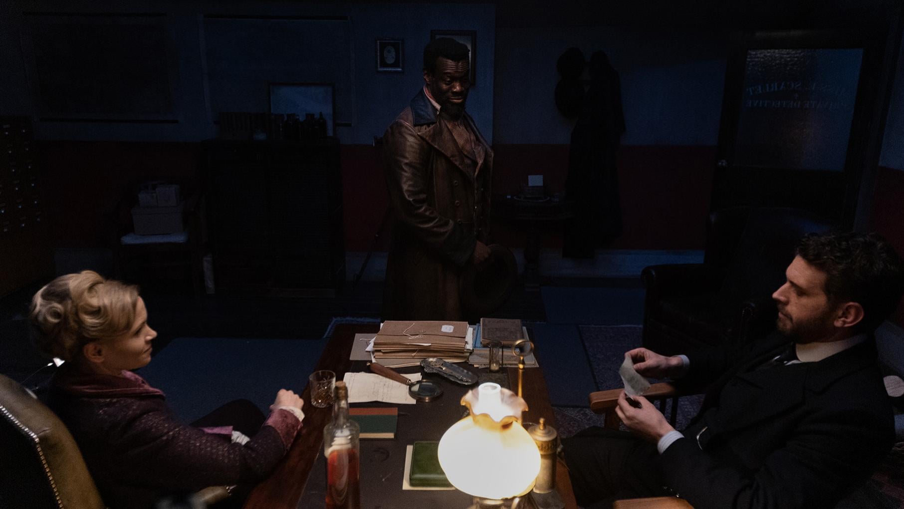 Eliza and Alexander sit across a desk from each other while Moses stands in the shadows of a dark room