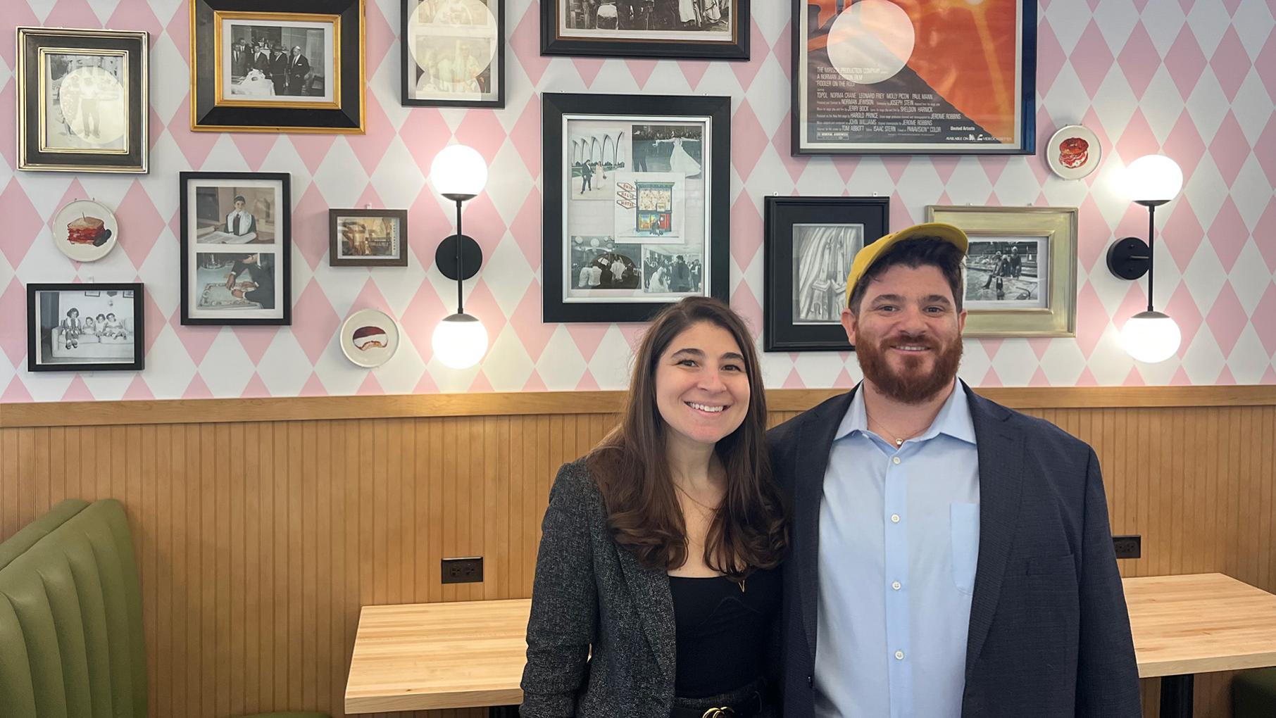 Ariel and Jake Schneider smile for a photo in their deli in front of a wall of family photos