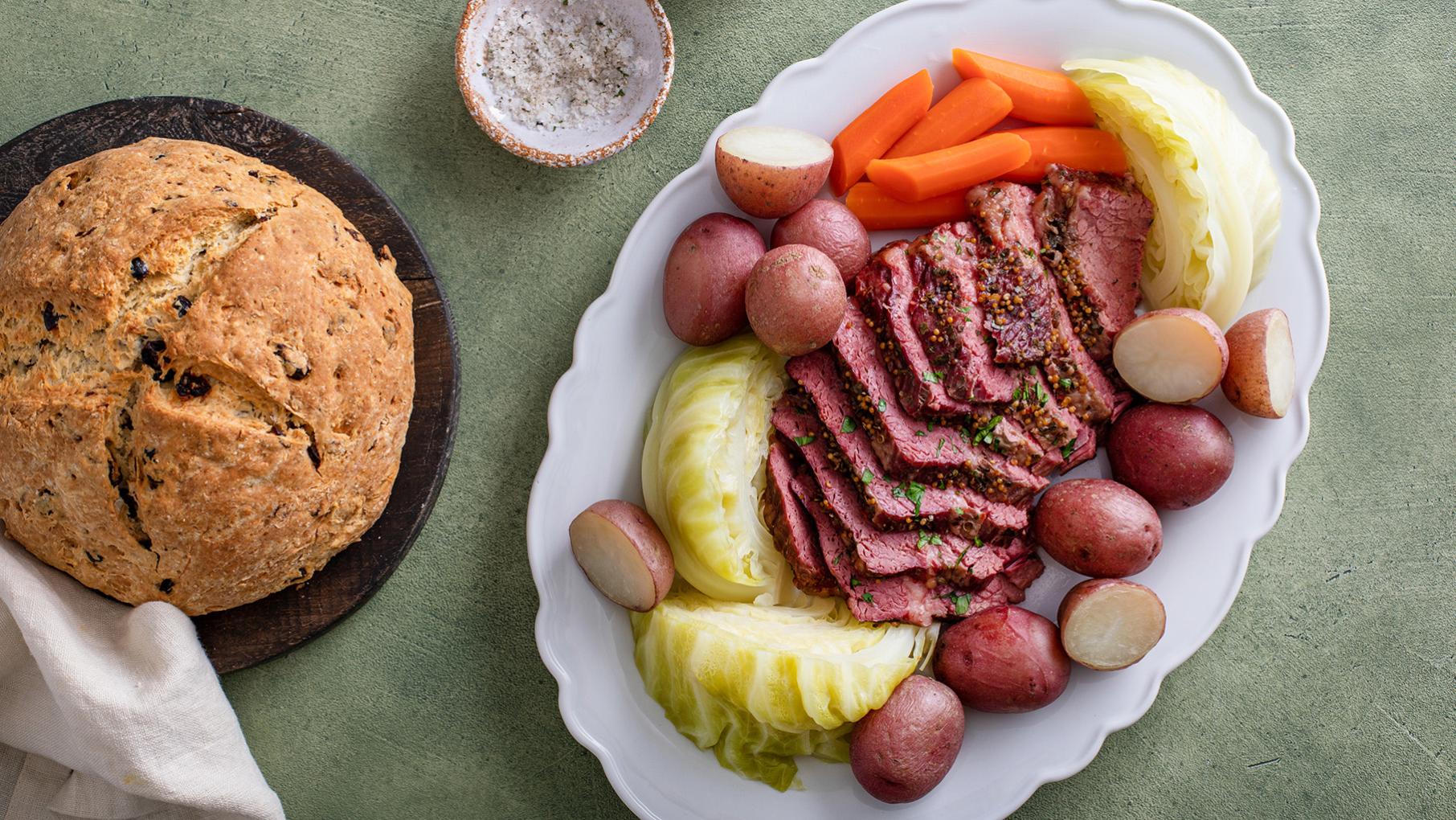 A platter of sliced corned beef, cabbage, potatoes, and carrots next to a loaf of soda bread