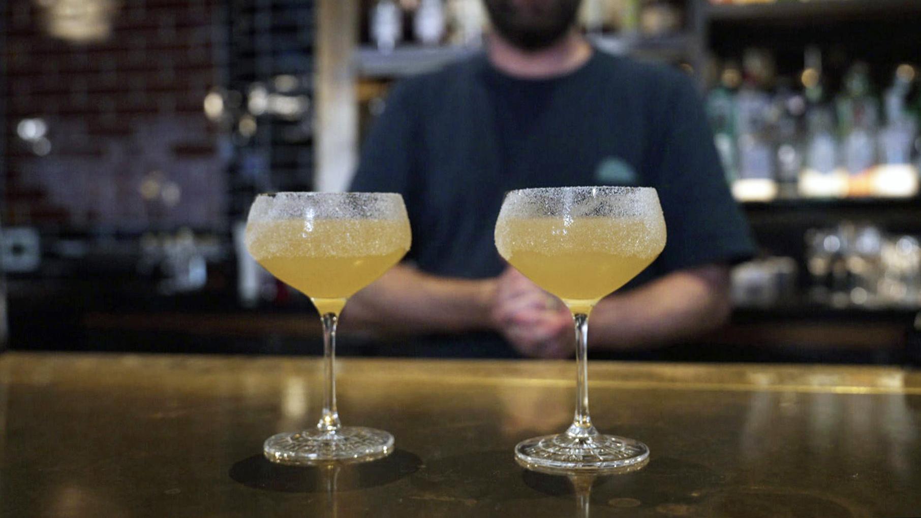 Two yellow cocktails in coupe glasses sit atop a bar while Liam Davy stands behind the bar out of focus