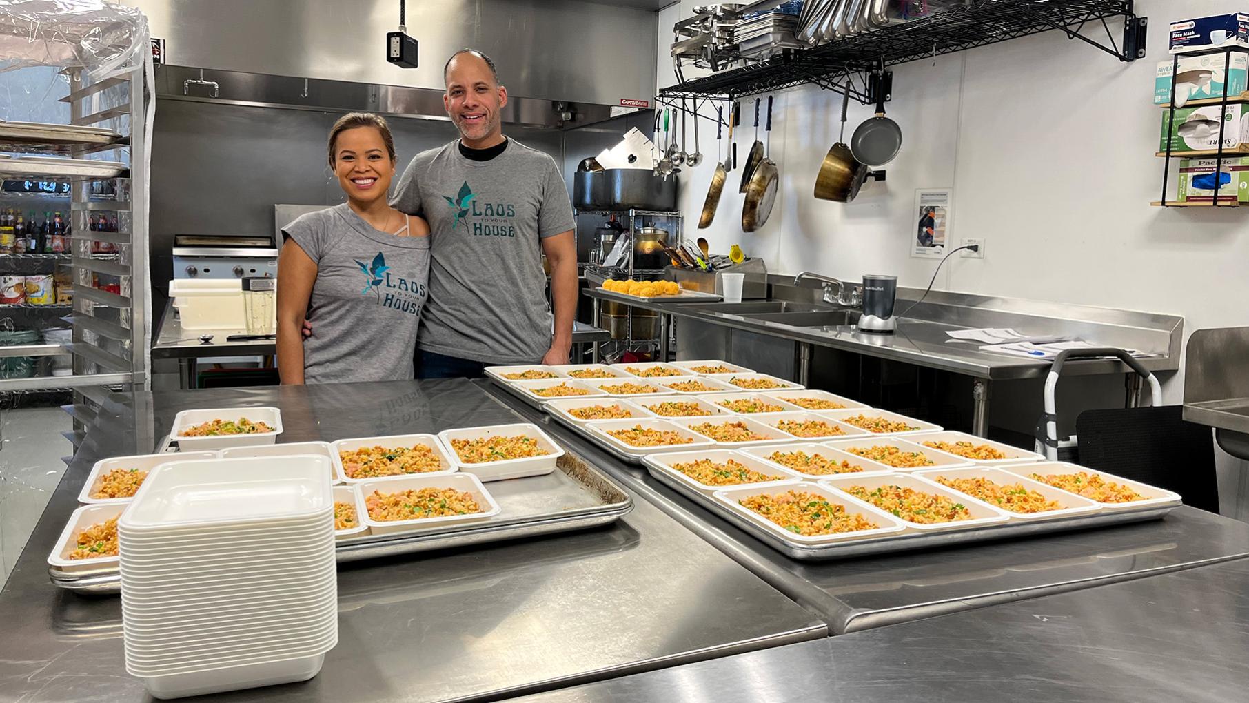 Two people in matching t-shirts pose in an industrial kitchen behind trays of food