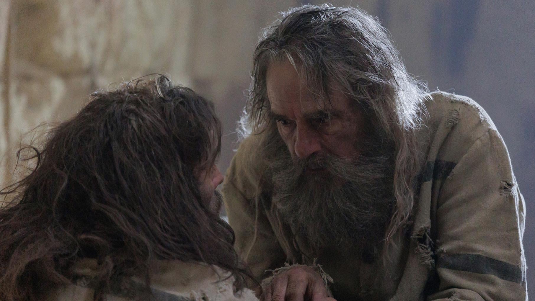 A heavily bearded man bends towards another, both in rags