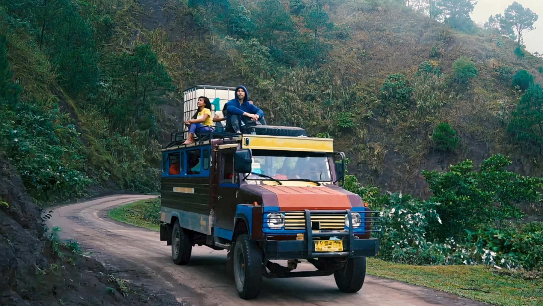 A colorful truck drives through verdant mountains on a road with two people sitting on top of it