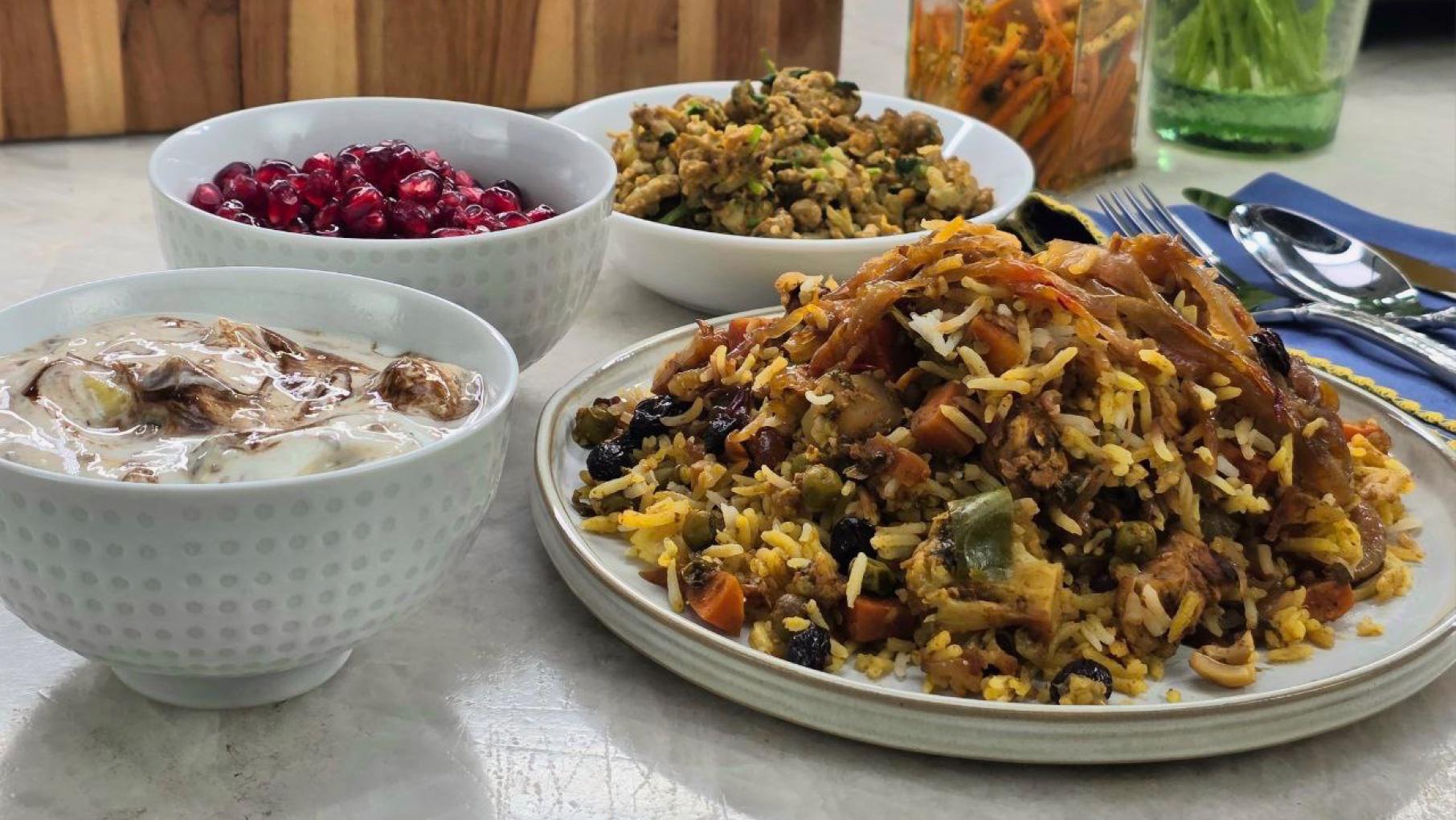 A plate of biryani next to a bowl of yogurt, pomegranate seeds, and another dish of food
