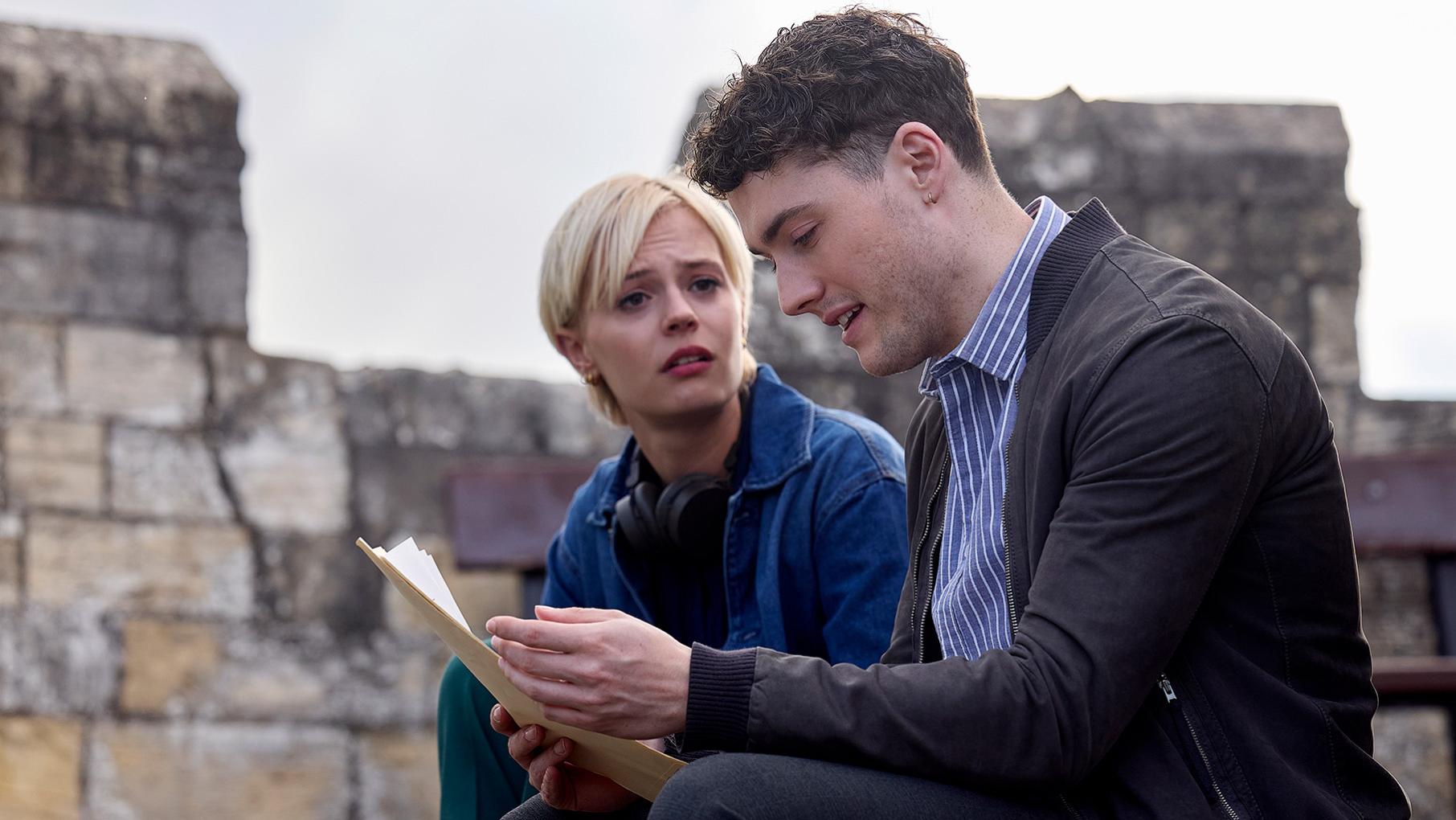 Patience looks concerned as Elliot reads a stack of papers on a medieval wall