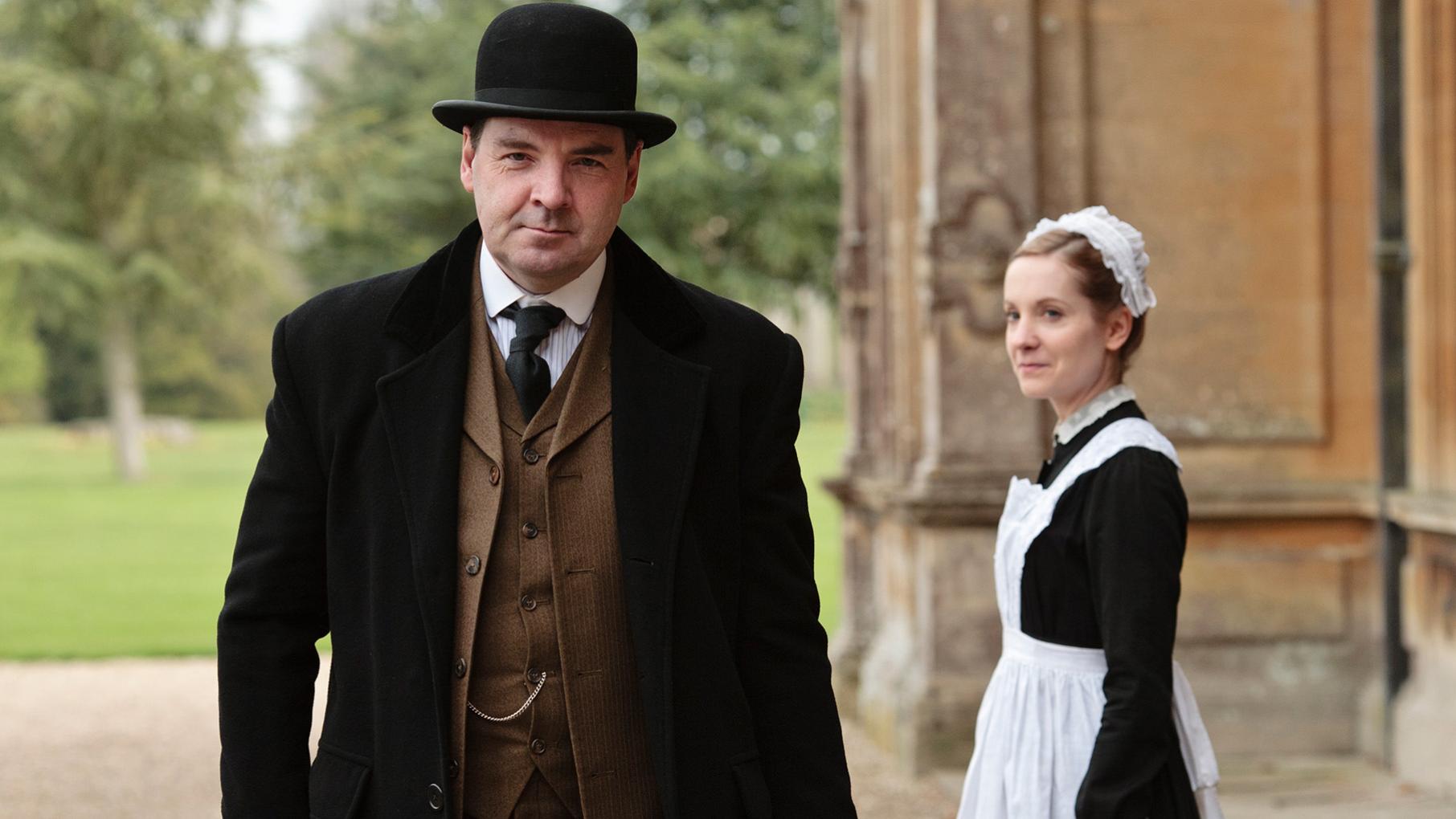 Bates walks in a jacket and hat past Anna outside