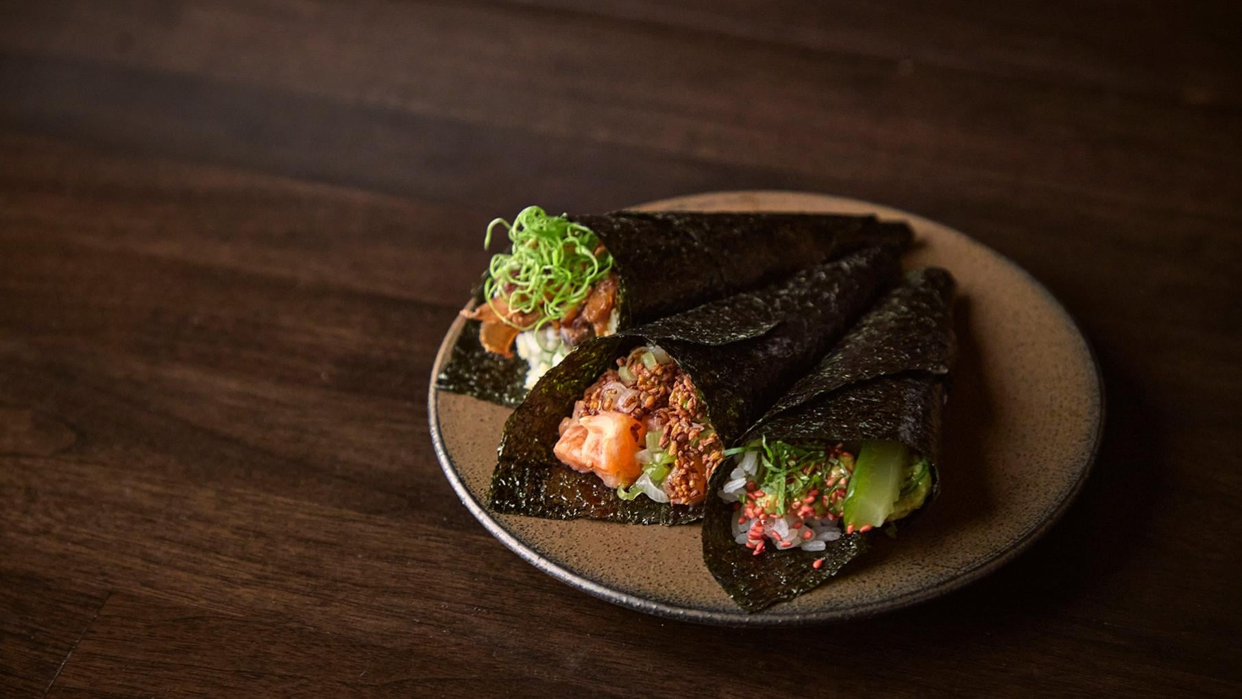 Three handrolls lie on a plate on a dark wood surface