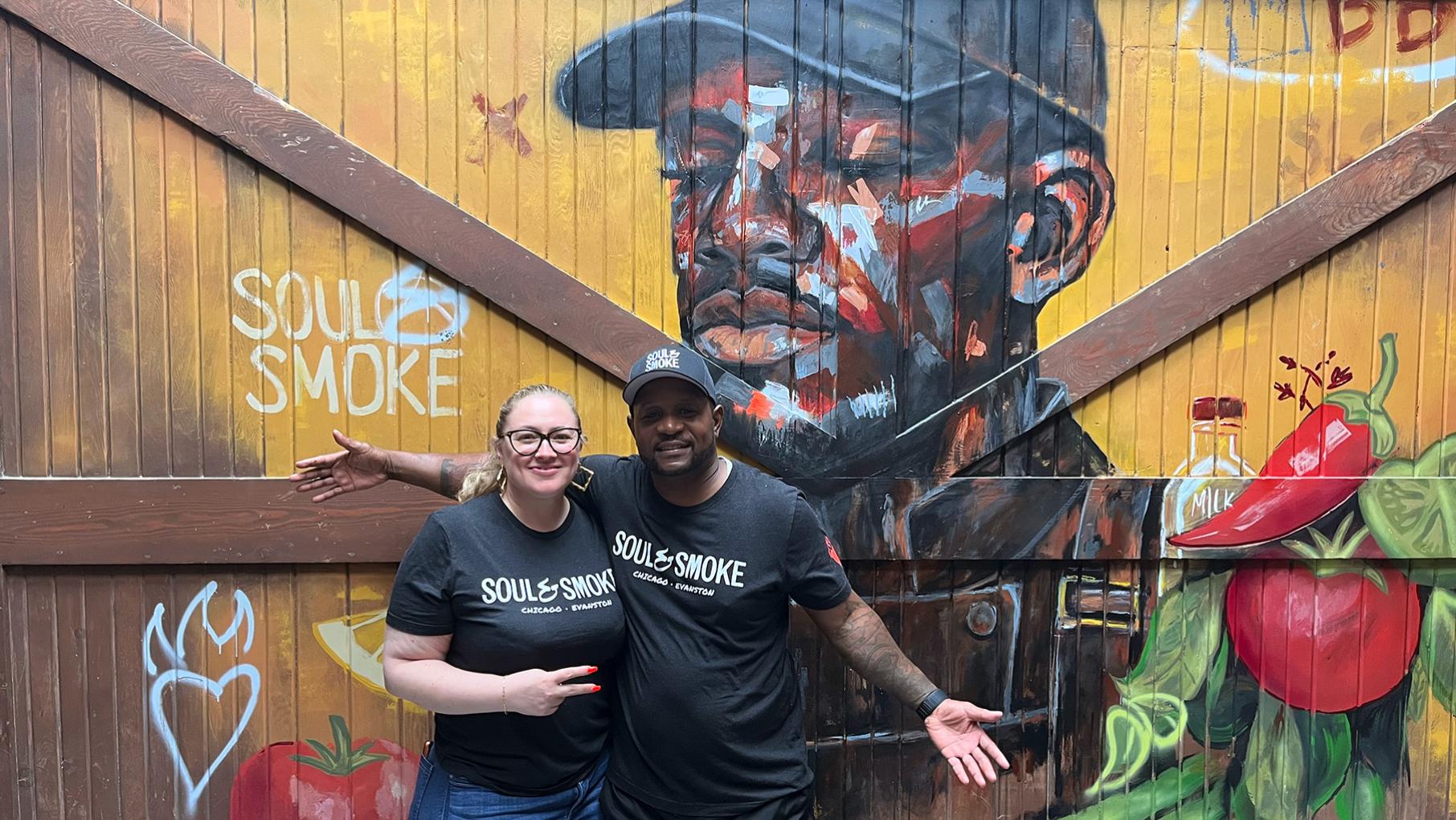 Heather Bublick and D'Andre Carter pose in front of a barn door painted with produce and a man in a cap