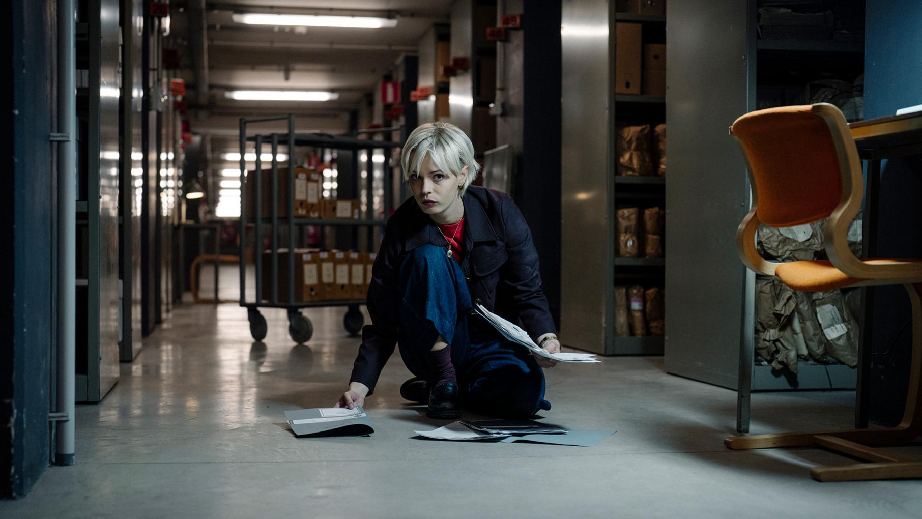 Patience squats to pick up papers from the ground in an archive