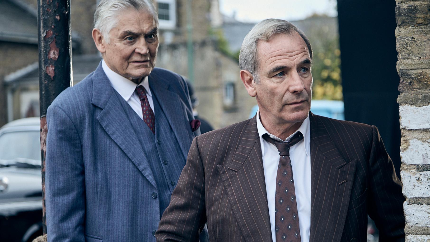 Jack and Geordie stand looking around the corner of a building in suits
