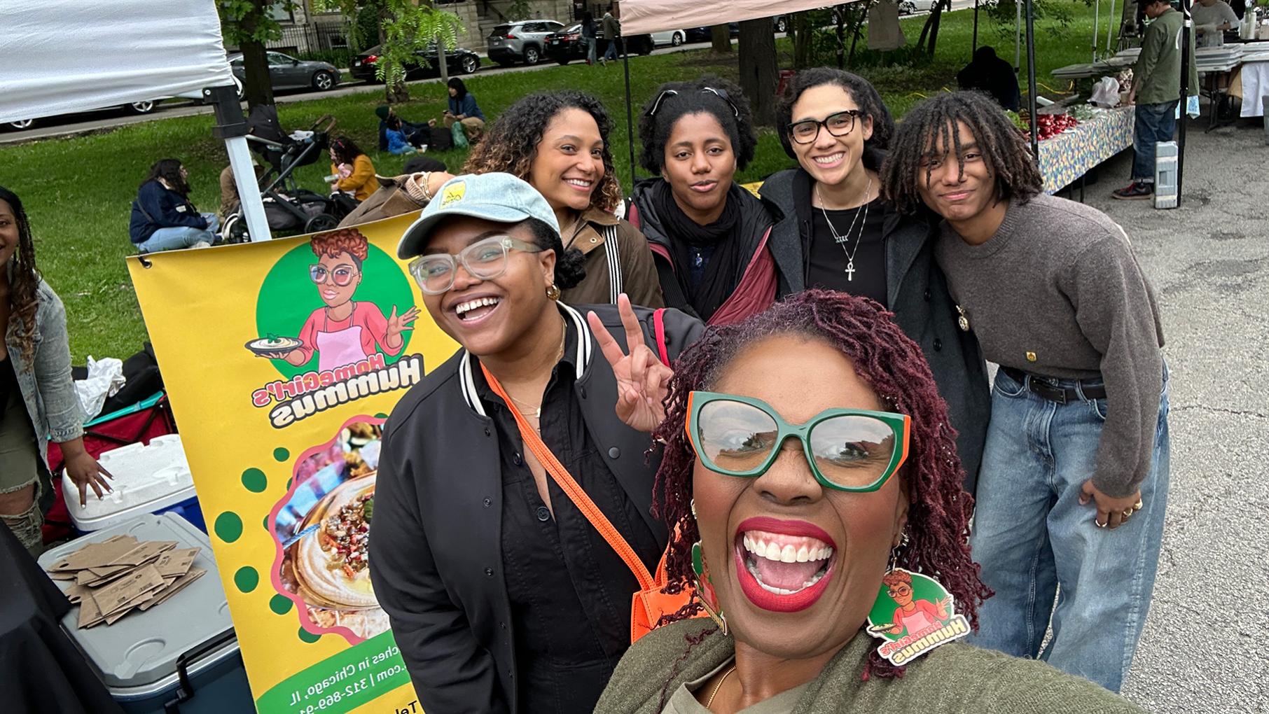 A woman takes a selfie of herself and a group behind her in front of a farmers market stand