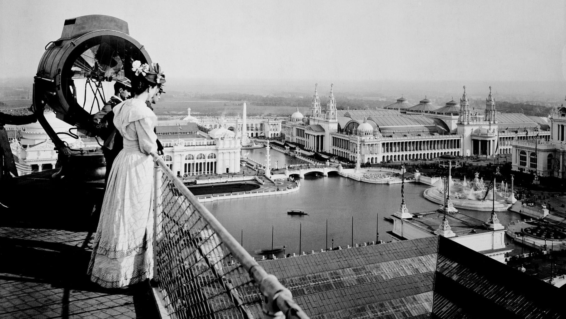 A black and white photo of some people looking over the 1893 World's Fair's White City and its lagoon from the roof of one of its buildings