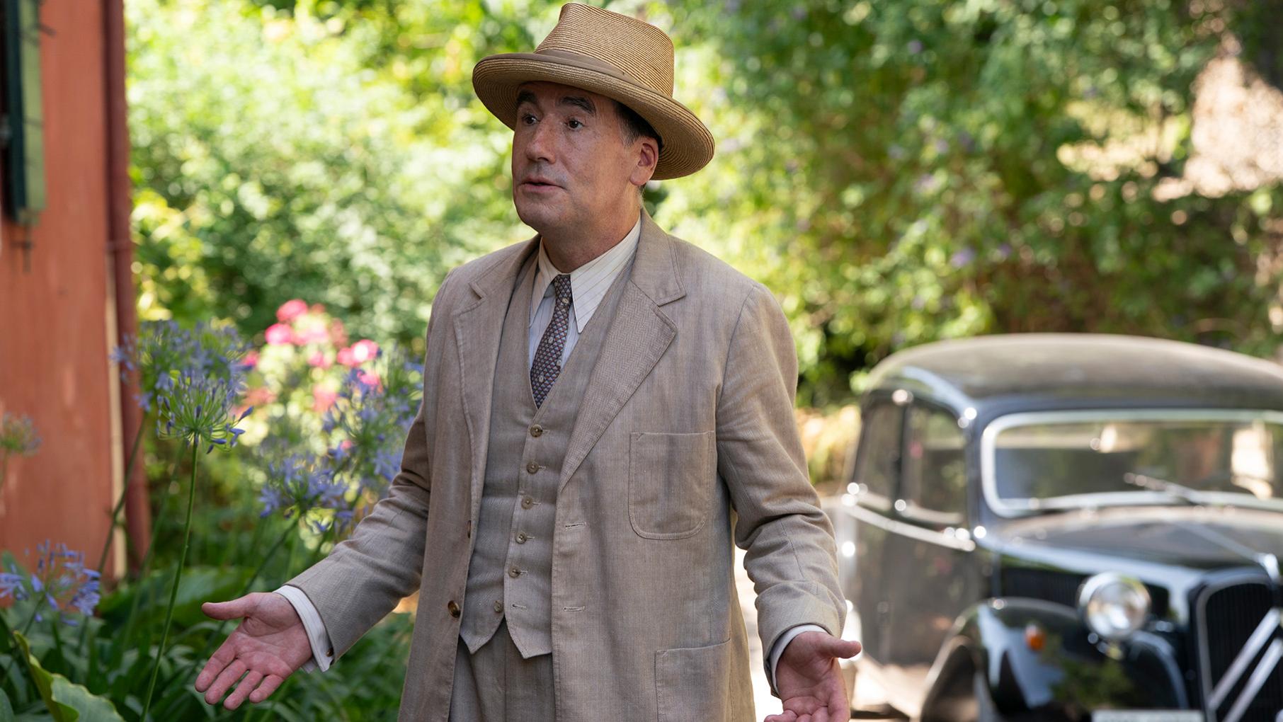 Atticus Pünd looks surprised standing outside in a hat and suit in front of a car