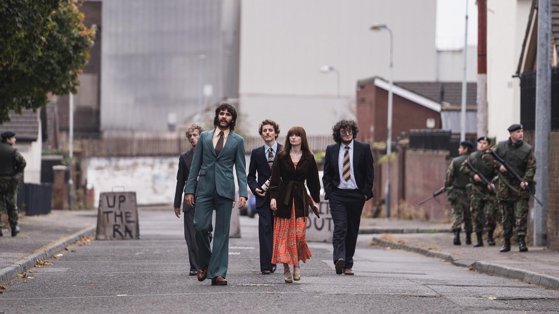 Gerry walks with Cushla and others down a street in nice clothes while soldiers stand on the sidewalk