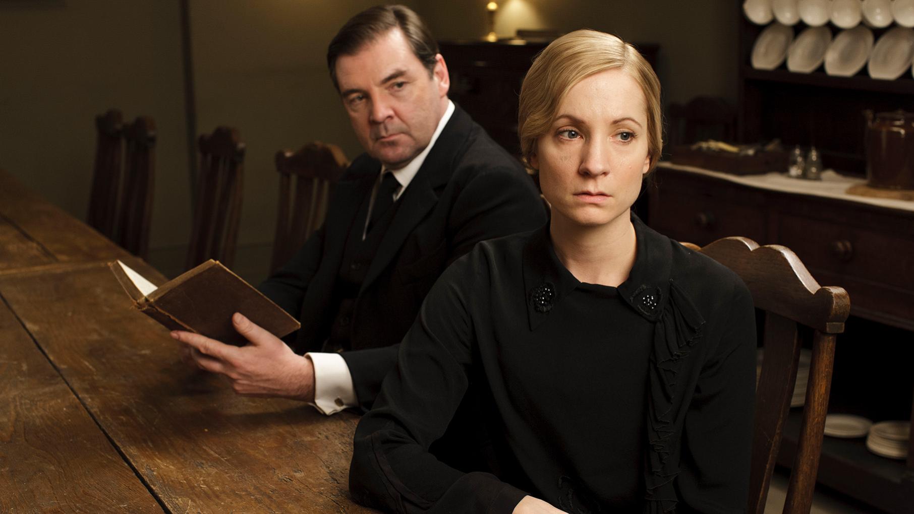 Anna looks morose in black while Bates sits behind her at a table holding a book