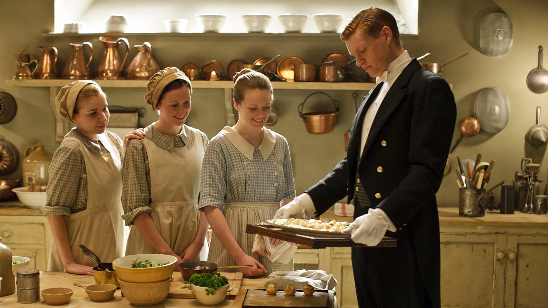 Alfred in a tuxedo and gloves holds a tray of food in the kitchen while three maids in aprons look on with smiles